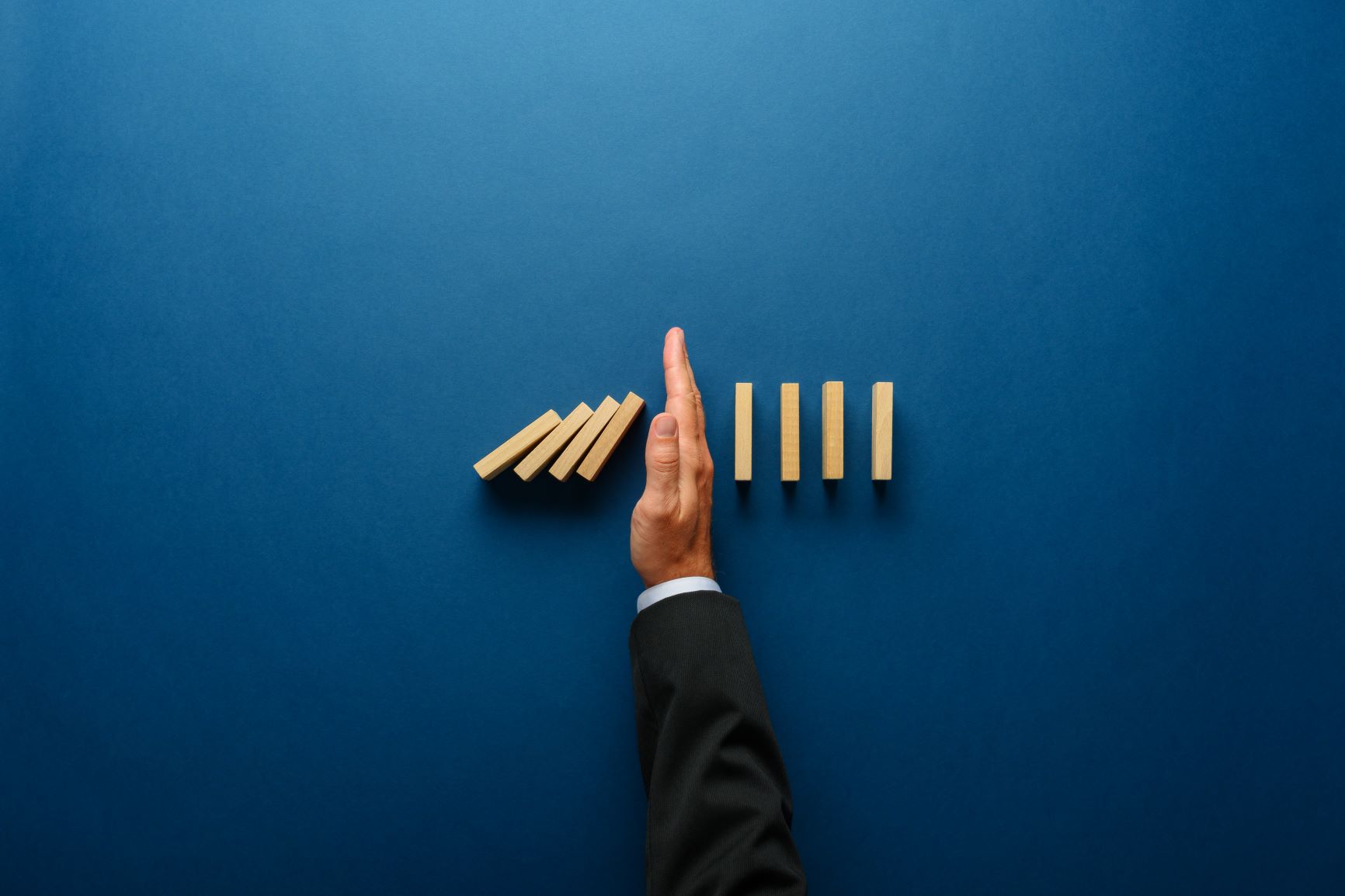 A hand in a suit stops a row of falling wooden dominoes on a blue background, preventing the remaining upright dominoes from toppling.