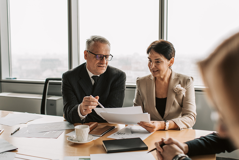 Two business professionals, a man in glasses and a woman in a beige blazer, sit at a conference table discussing documents. They are in a modern office with large windows and papers spread on the table.