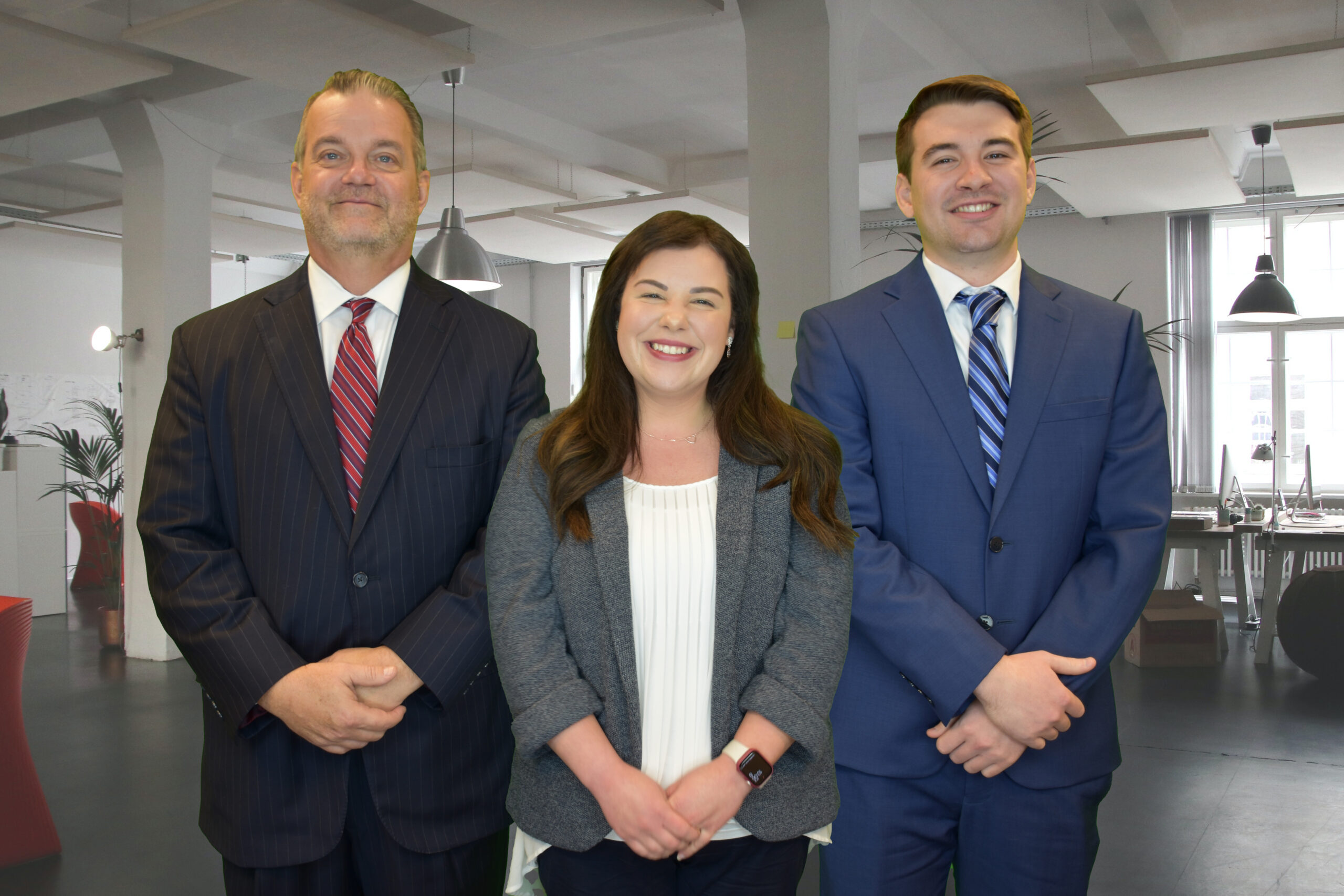 Three people in business attire—two men in suits and a woman in a blazer—stand smiling in a modern, bright office space with large windows and desks in the background.