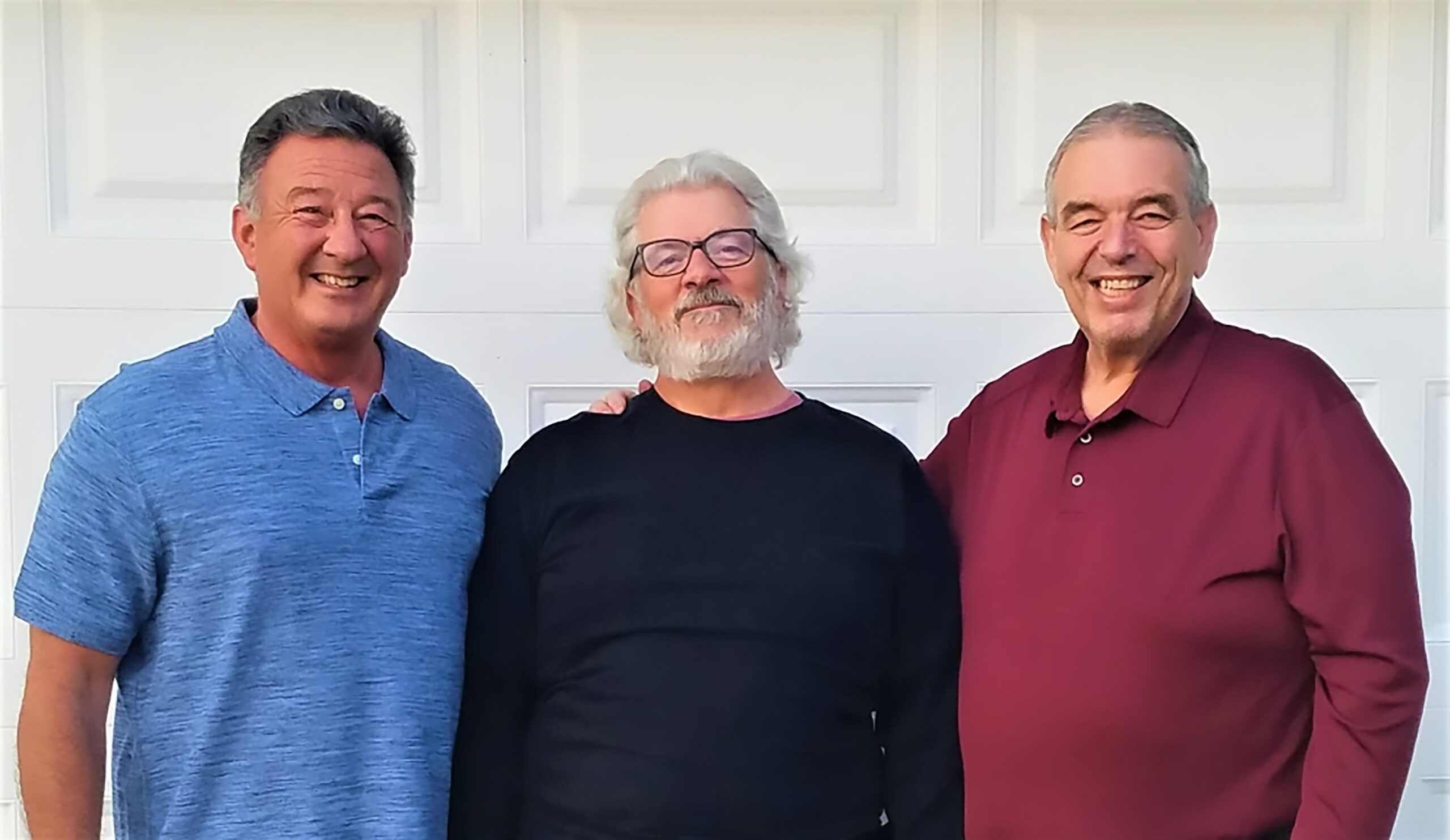 Three older men stand close together in front of a white garage door, smiling at the camera. The man on the left wears a blue shirt, the center man has glasses and a black shirt, and the man on the right wears a maroon shirt.