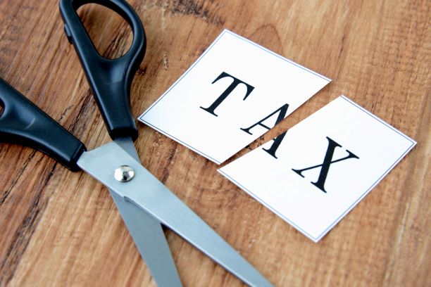 A pair of scissors cutting a piece of paper with the word TAX printed on it, symbolizing a tax cut or reduction, on a wooden surface.