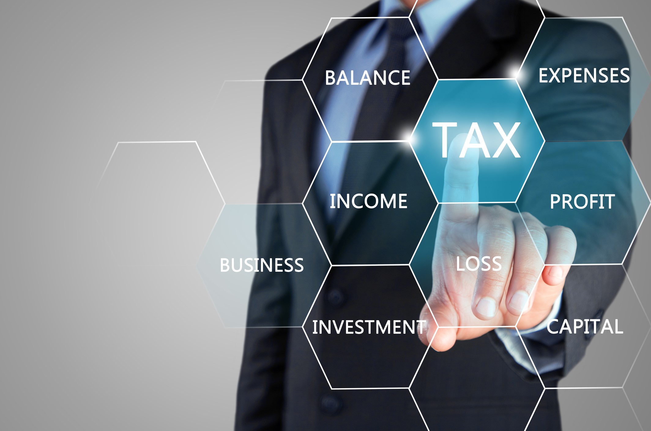 A person in a suit touches a virtual hexagonal button labeled TAX among other financial terms like income, expenses, profit, loss, investment, balance, business, and capital.