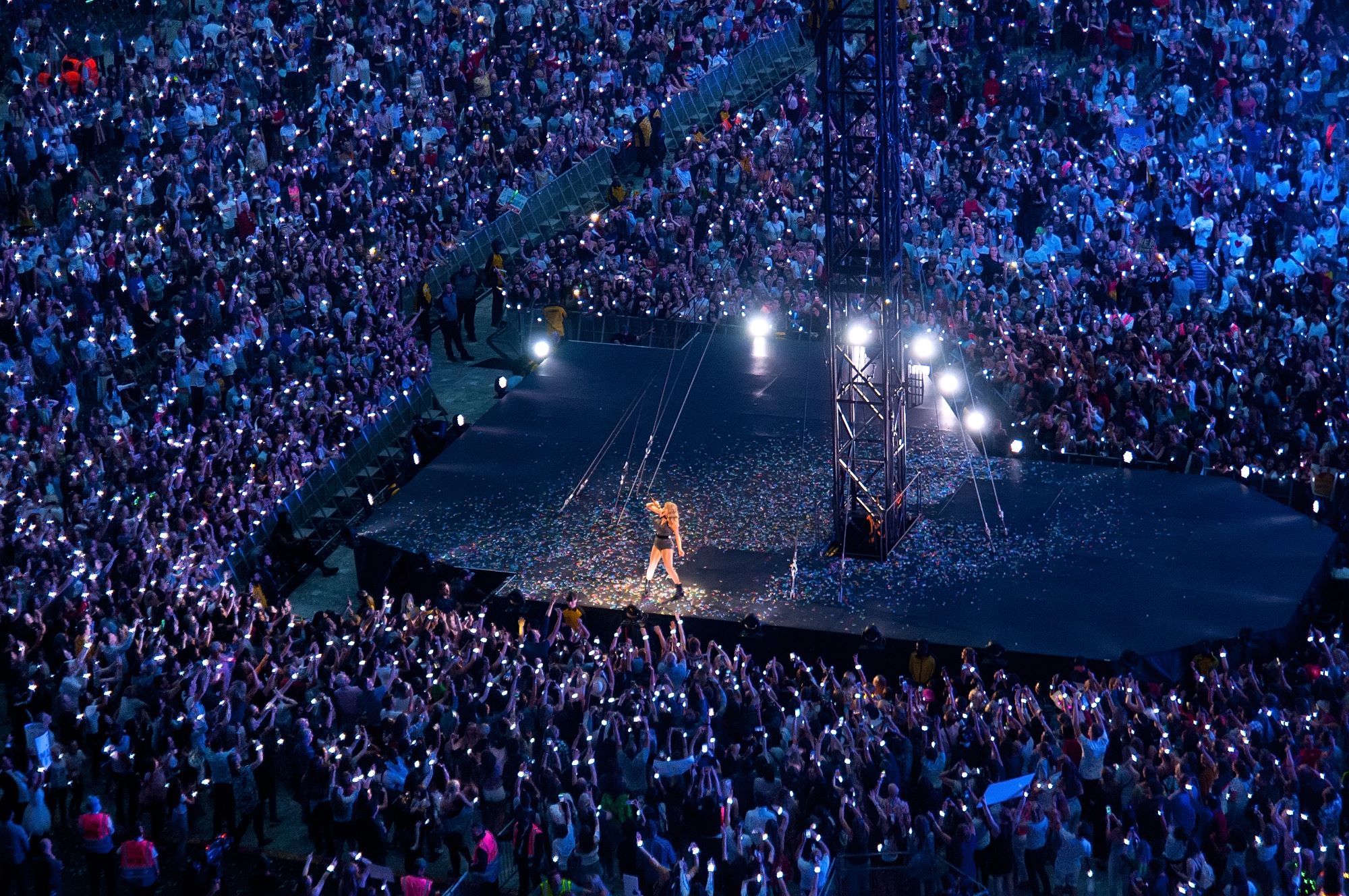 A performer stands on a large stage in the center of a packed stadium, surrounded by thousands of fans holding up glowing lights, creating a sea of illuminated spectators at a nighttime concert.