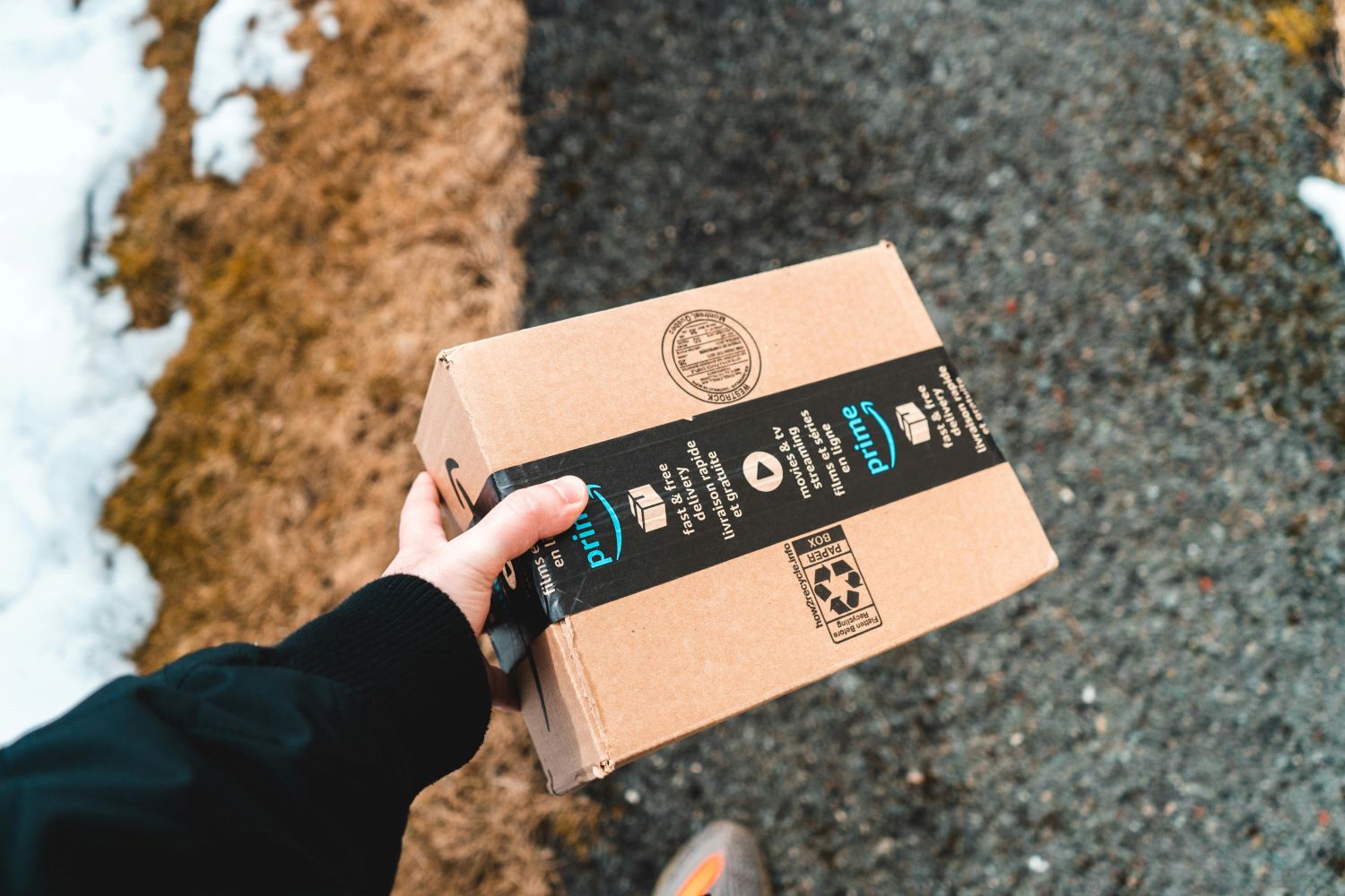 A person’s hand holding a small Amazon Prime package outdoors on a gravel path, with patches of snow and dry grass visible on either side.