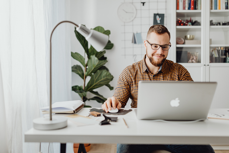 A man with glasses and a beard sits at a desk, smiling while working on a laptop. The desk has an open notebook, documents, and a lamp. There are plants and shelves in the well-lit, modern room.