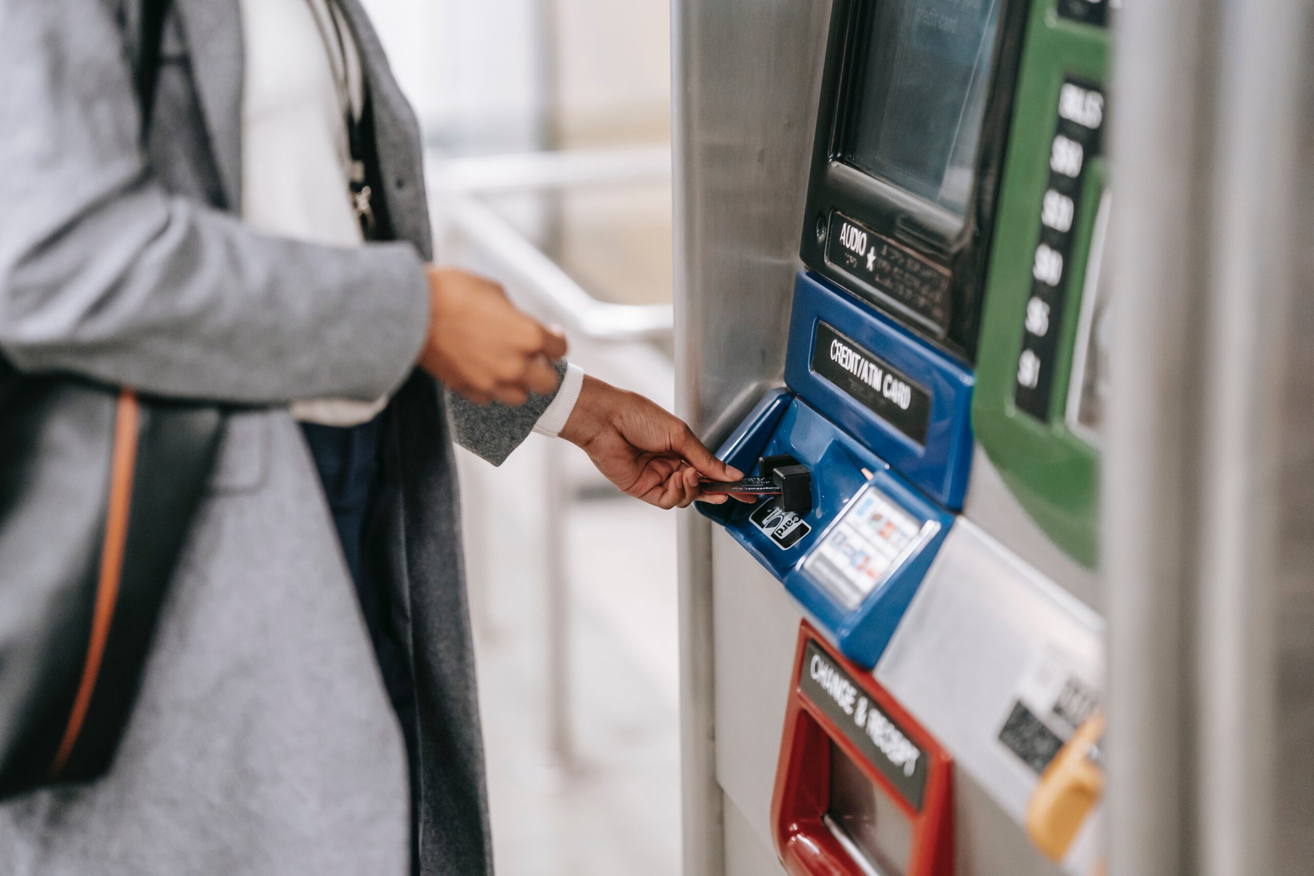 A person wearing a gray coat inserts a card into a fare payment machine, possibly at a subway or train station. The machine has buttons labeled for credit card and change options.
