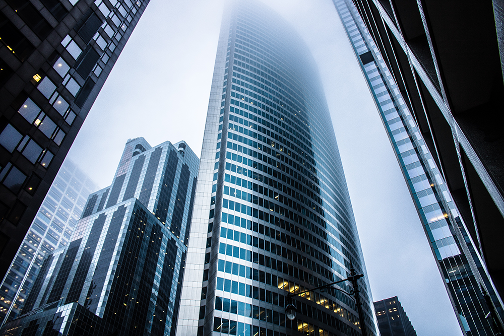 Tall modern skyscrapers rise into a foggy sky, their glass and steel exteriors reflecting light. The tops of the buildings are partially obscured by mist, creating a dramatic and atmospheric city scene.