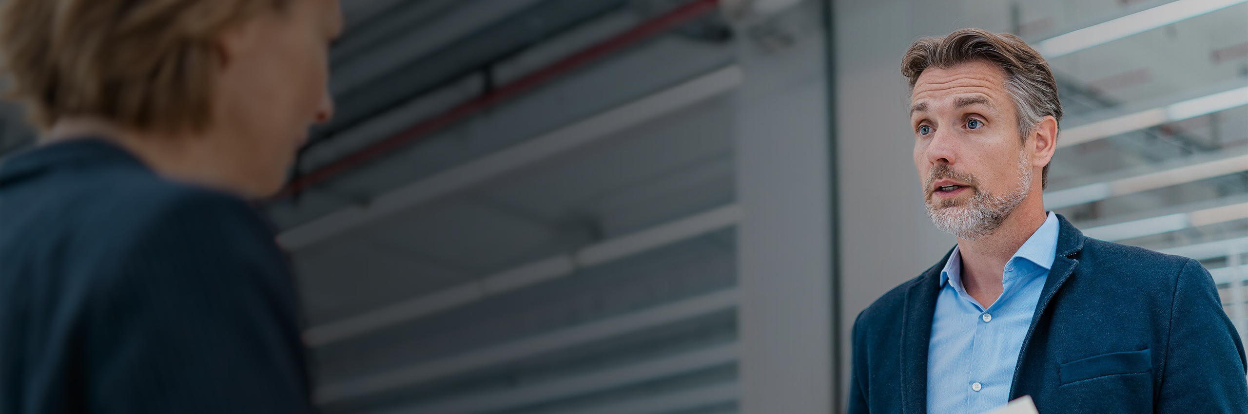A man with gray hair and a beard, wearing a blue blazer and shirt, talks to a person whose back is to the camera in a modern office setting.