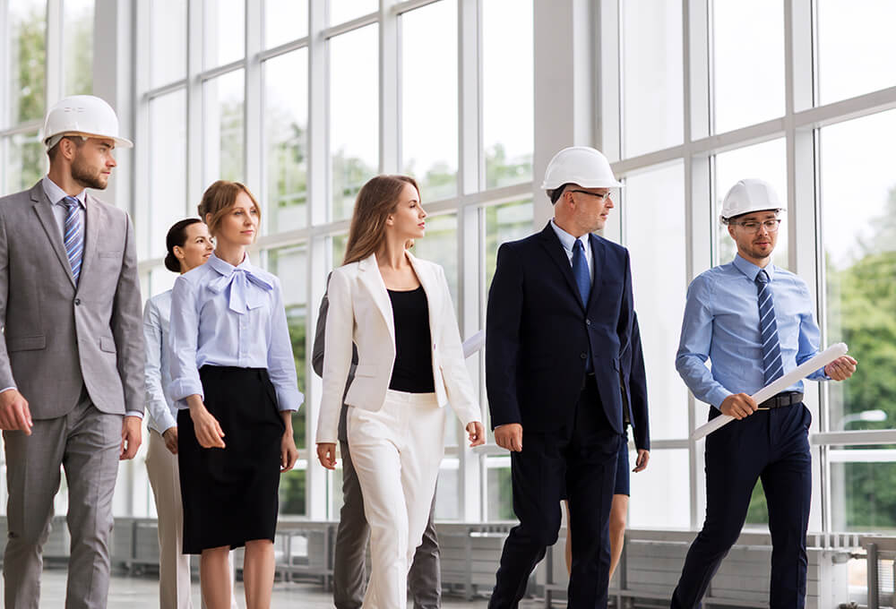 A group of business professionals, some wearing hard hats, walk together in a modern building with large windows. One person holds blueprints, suggesting a meeting or site inspection.