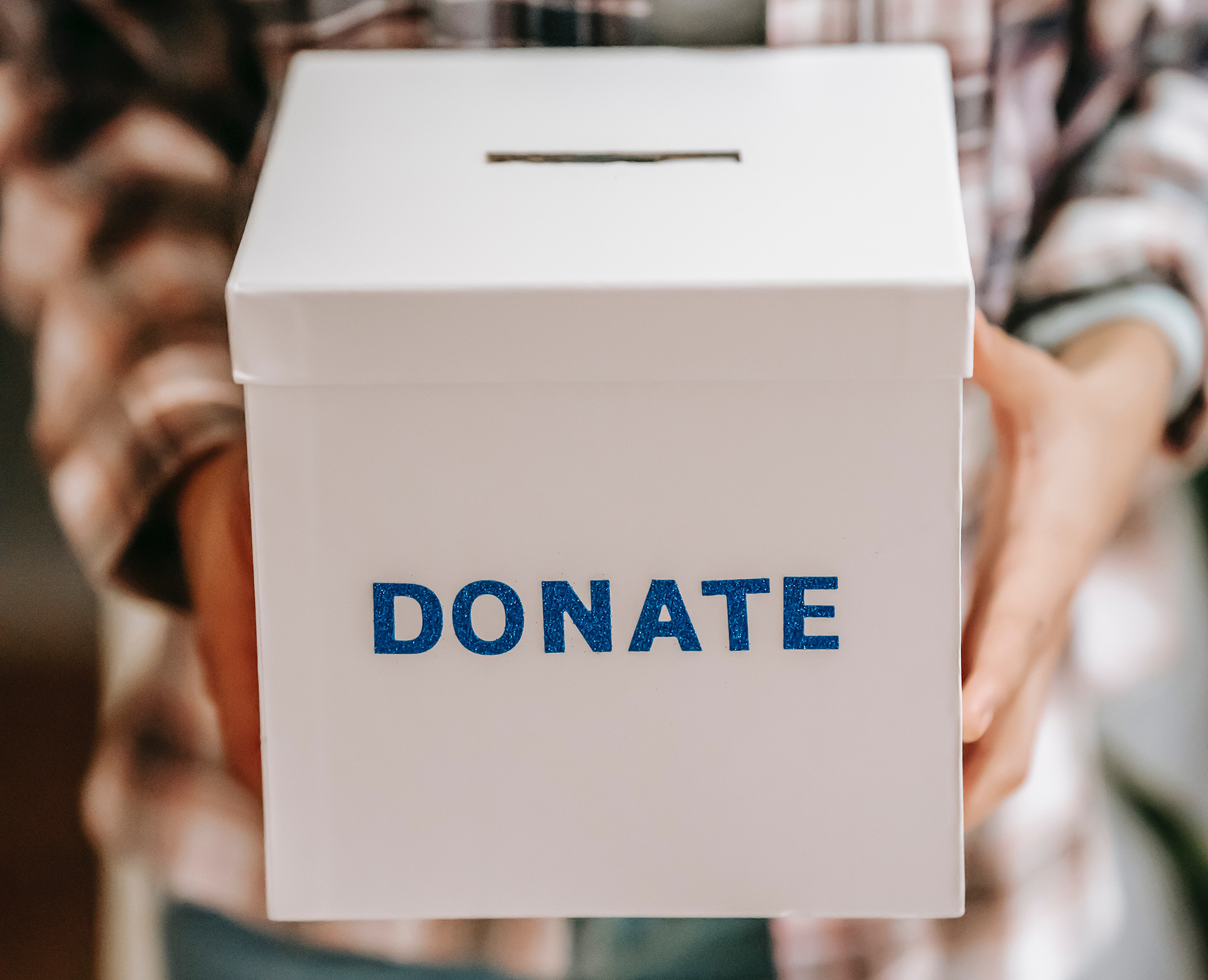 A person holding a white donation box with a slot on top and the word DONATE written in blue letters on the front.