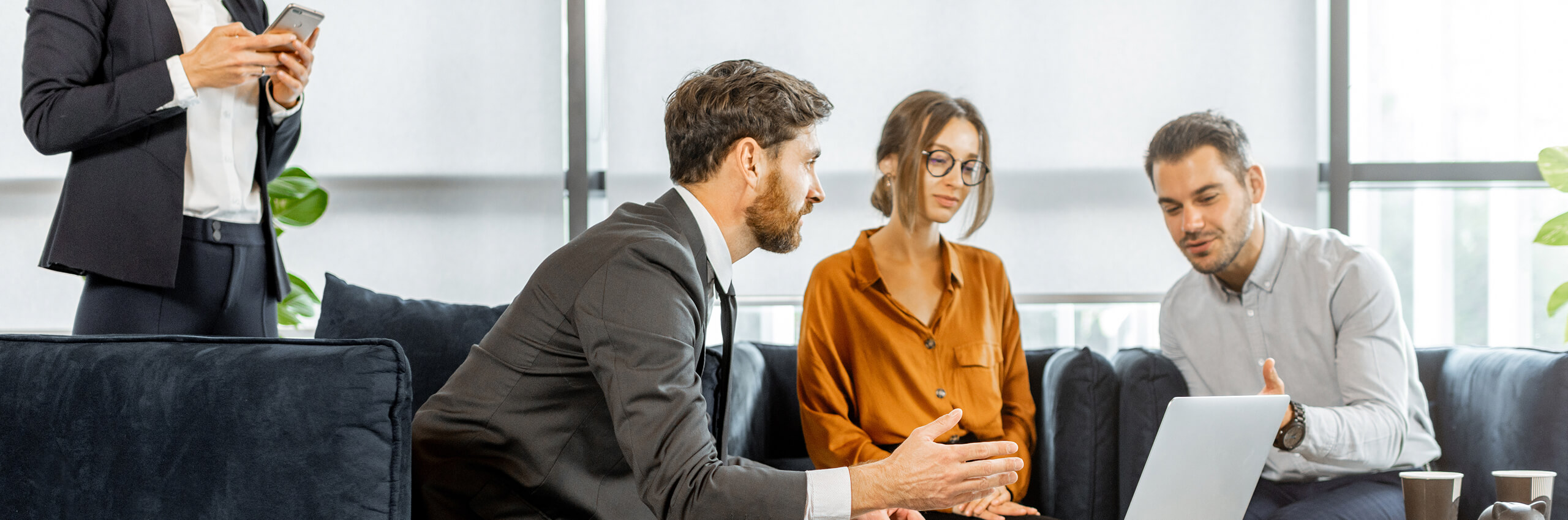 Three people sit on couches discussing something while looking at a laptop, with another person standing nearby using a smartphone. The setting appears to be a modern office or lounge area.
