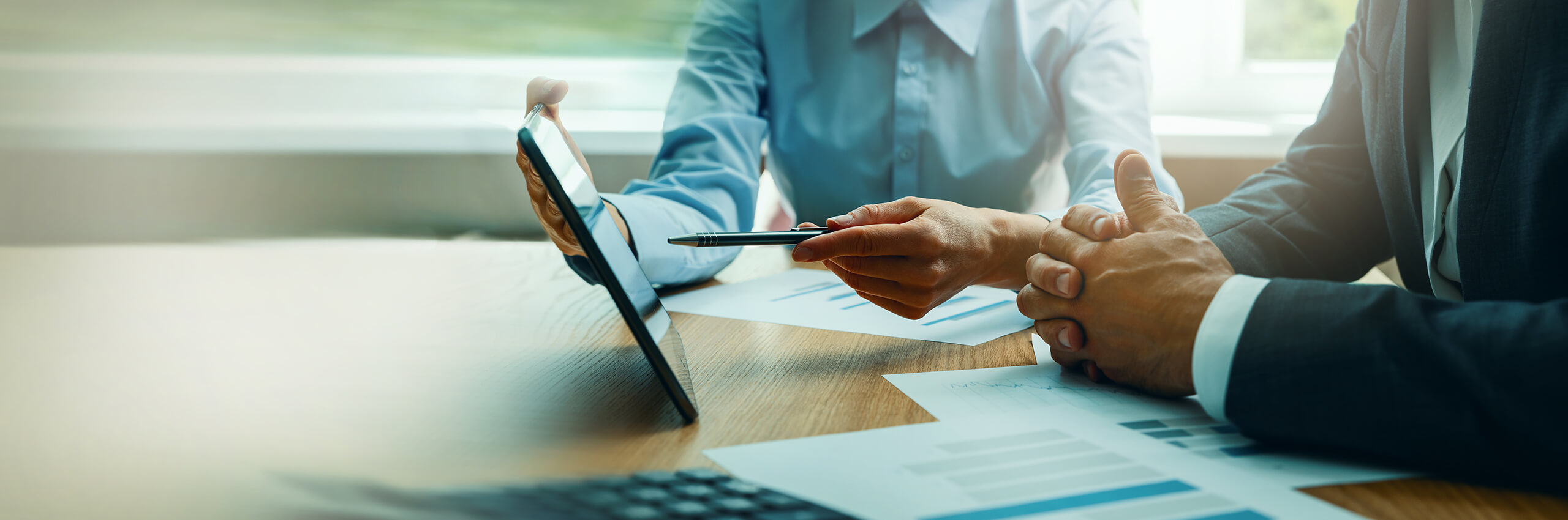 Two people in business attire sit at a desk, discussing a document on a tablet. One person gestures with a pen, while charts and papers are spread on the table in front of them.
