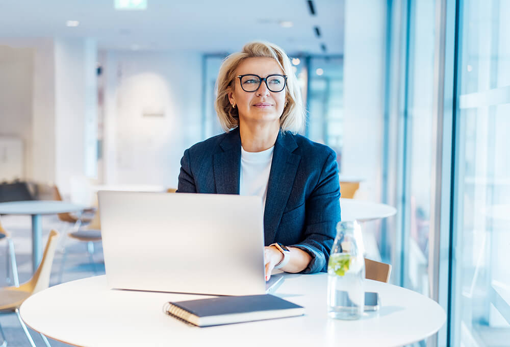 A woman in business attire sits at a table with a laptop, notebook, and a glass of water. She is looking thoughtfully out the window in a bright, modern office or cafe.