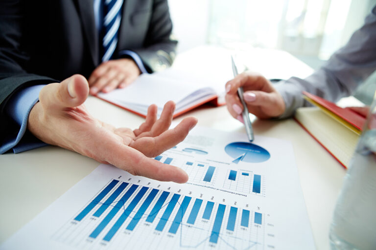 Two people in business attire discuss financial charts and graphs at a desk, one gesturing with their hand while the other points with a pen. A bar graph and pie chart are visible on a printed report in the foreground.