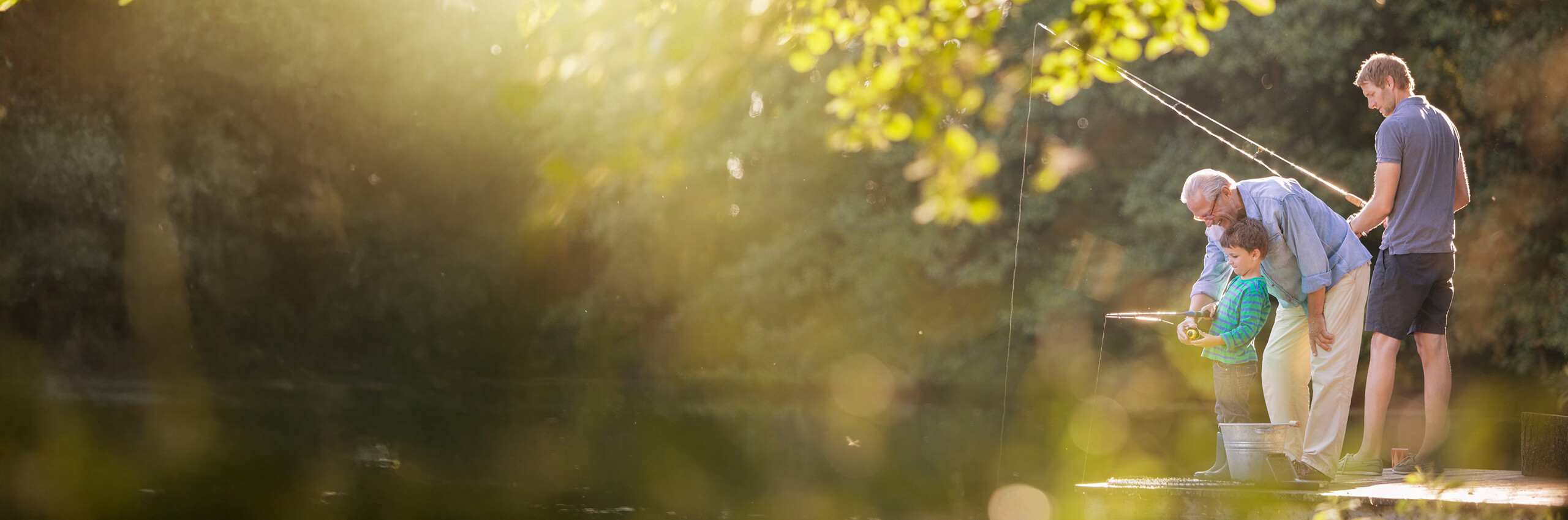 Three generations—an elderly man, a young boy, and a younger man—stand on a dock fishing together, surrounded by sunlight and greenery, creating a warm, peaceful outdoor scene.