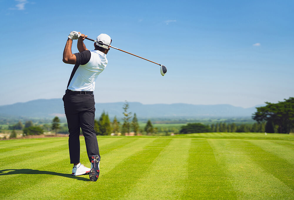 A golfer is swinging a club on a neatly mowed golf course, facing away from the camera. The sky is clear, and mountains and trees can be seen in the distance.