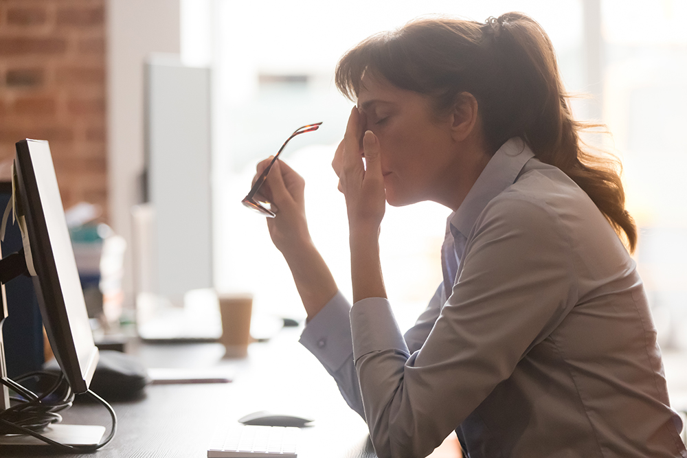 A woman sits at a desk in front of a computer, holding her glasses in one hand and pinching the bridge of her nose with the other, appearing tired or stressed.