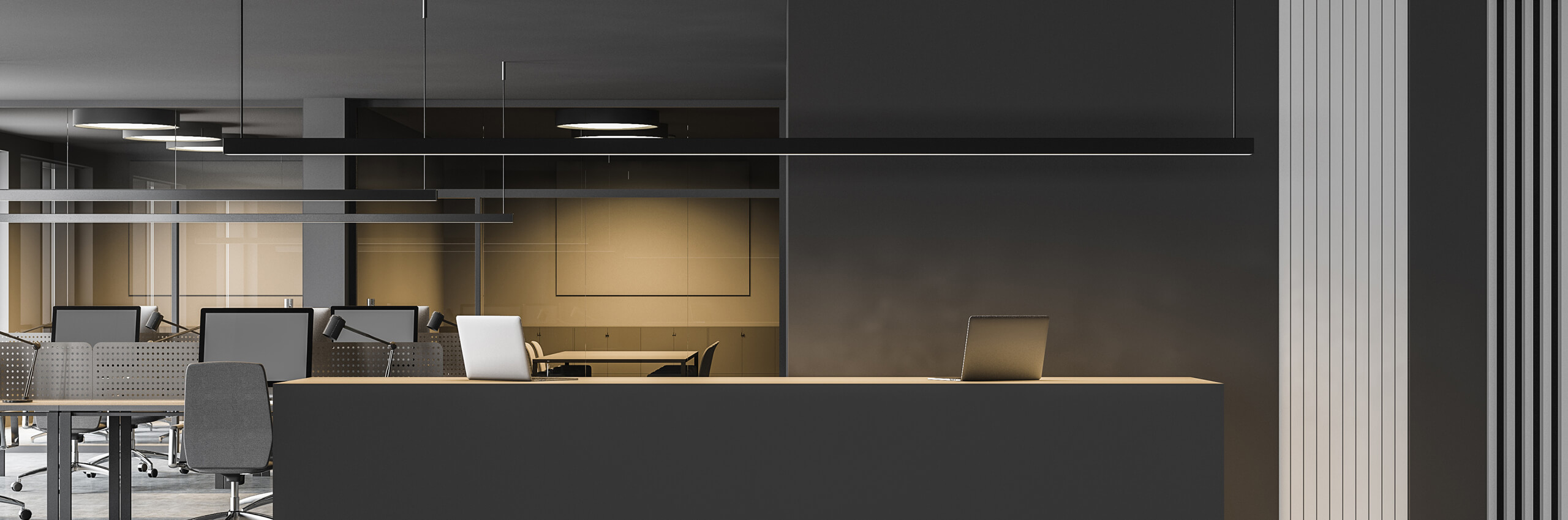 A modern office space with minimalist black and gray décor, featuring a front desk with a laptop, several office chairs, desks with monitors, and hanging ceiling lights.
