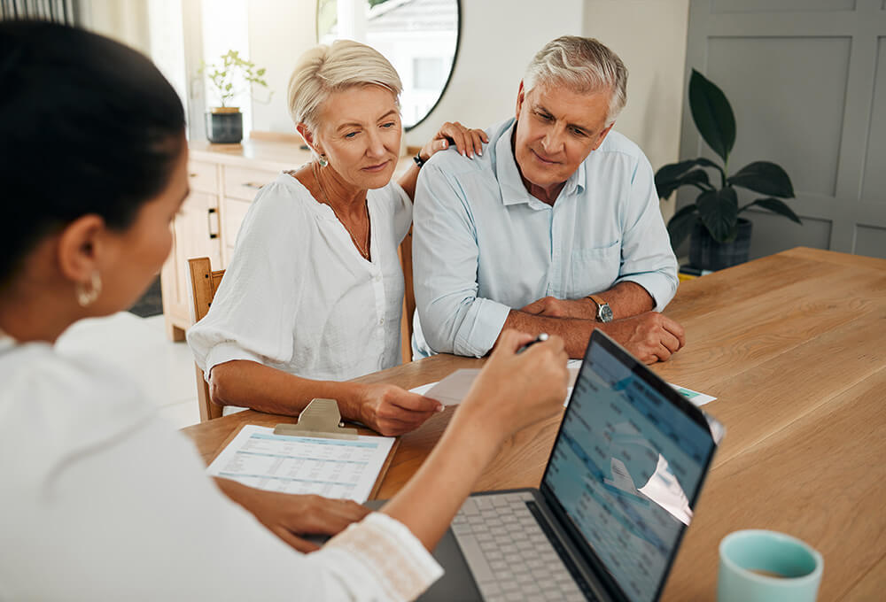 An older couple sits at a table, attentively discussing documents with a professional who is showing financial information on a laptop. The atmosphere is calm and focused.