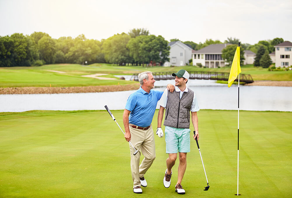 An older and a younger man, both holding golf clubs, walk together on a golf course near a flag. They smile at each other, with houses, trees, and a pond visible in the background.