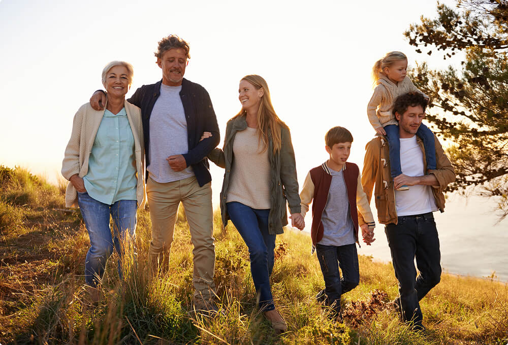 A multi-generational family of six walks outdoors on a grassy hill at sunset. Two adults, two children, a young woman, and an older woman appear happy, with one child riding on an adult’s shoulders.