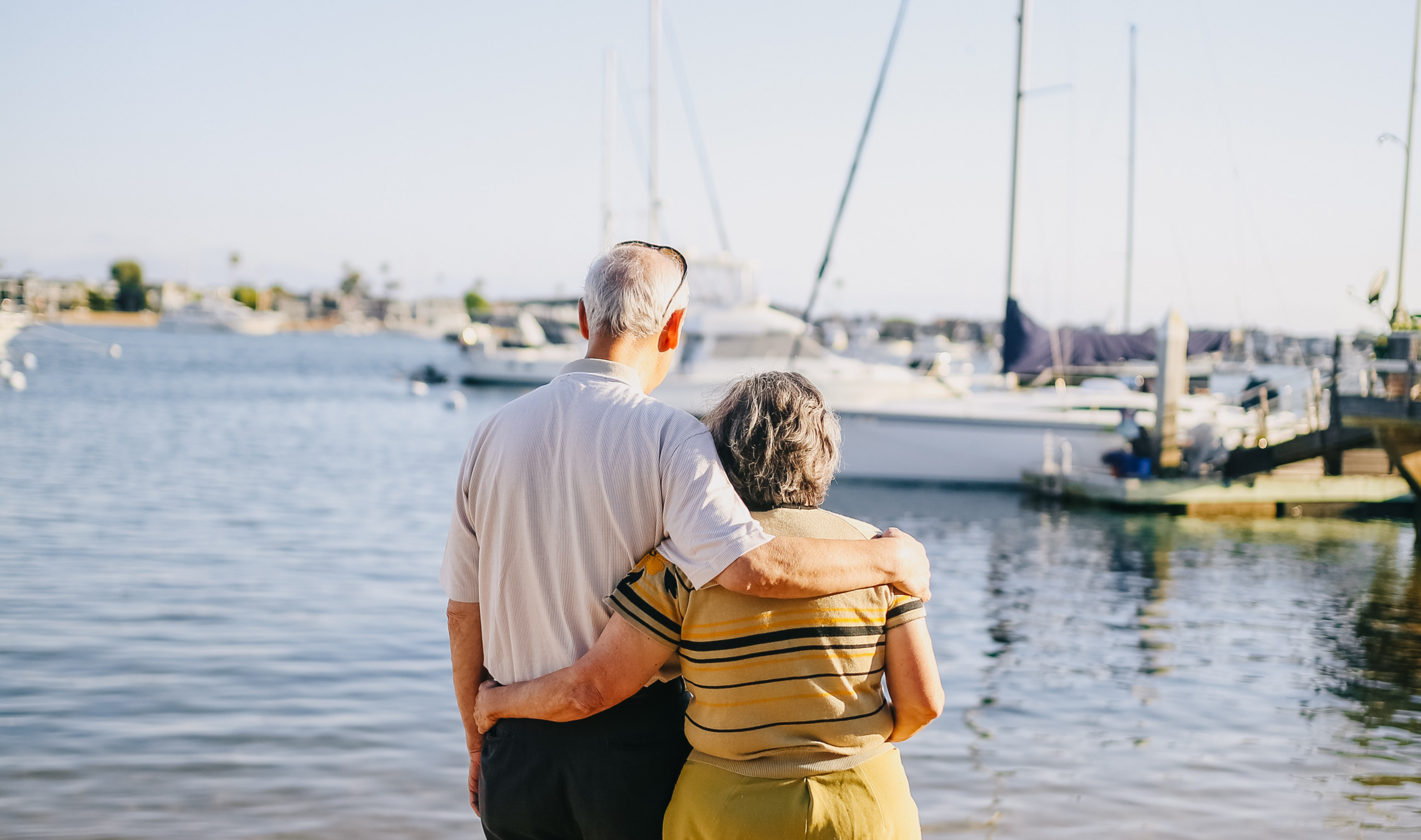 An elderly couple stands arm in arm by a marina, looking out at sailboats docked on calm water under a clear blue sky.