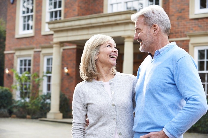 Smiling older couple stands close together outside a large brick house, looking at each other happily. Both wear light sweaters, and the scene appears bright and cheerful.