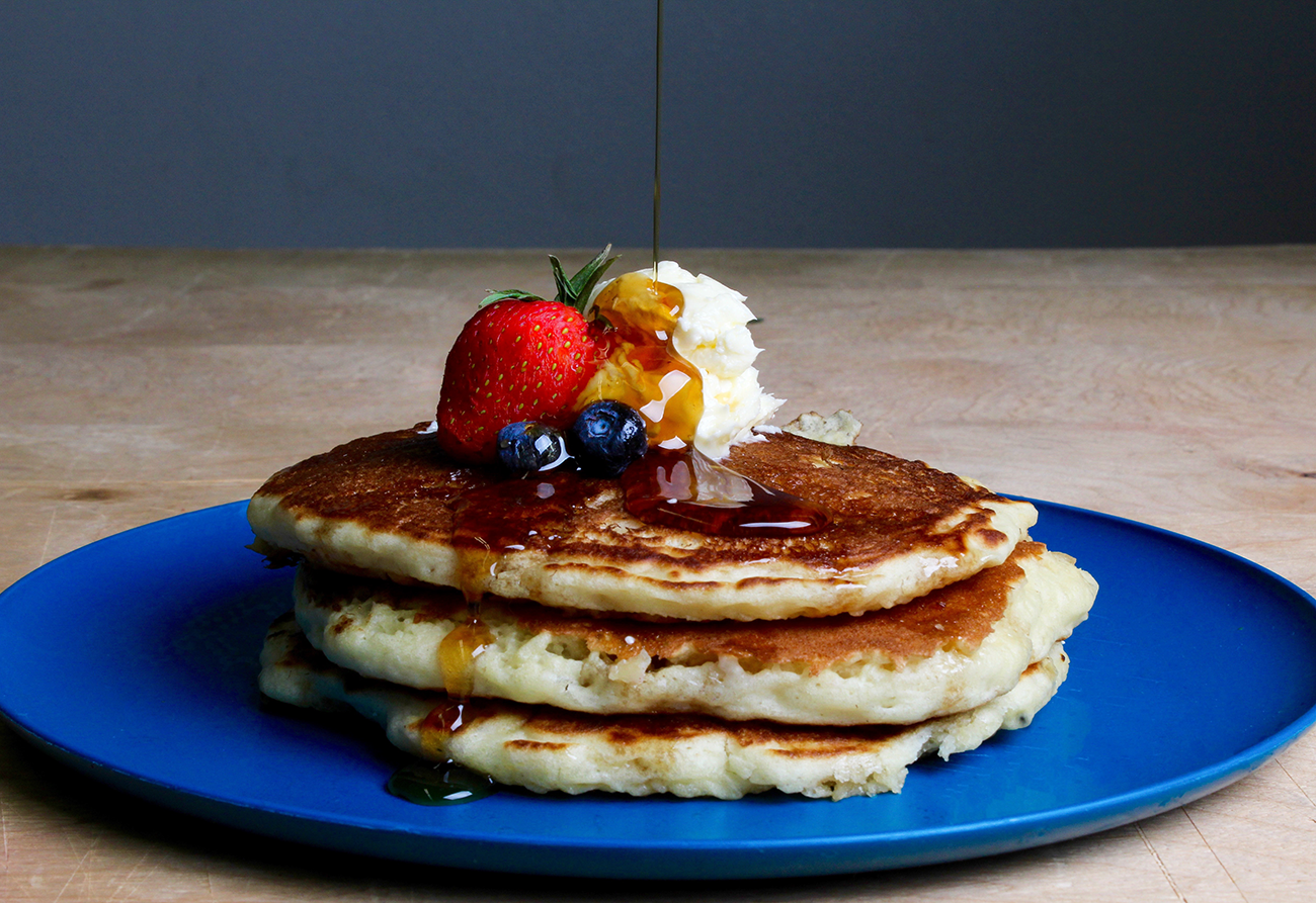 Three fluffy pancakes stacked on a blue plate, topped with whipped cream, a strawberry, and blueberries, with syrup being poured over them. The background is a plain wooden surface and a gray wall.