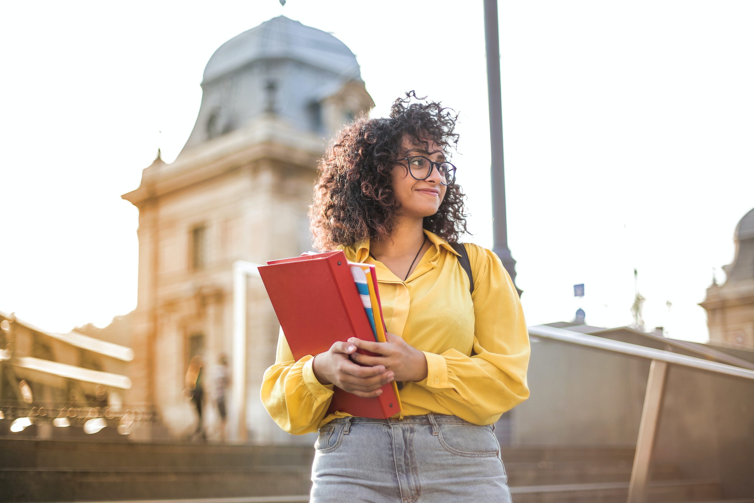 A young woman with curly hair, wearing glasses and a yellow shirt, stands outdoors holding red folders and notebooks. There is a historic building and stairs in the background, with sunlight illuminating the scene.