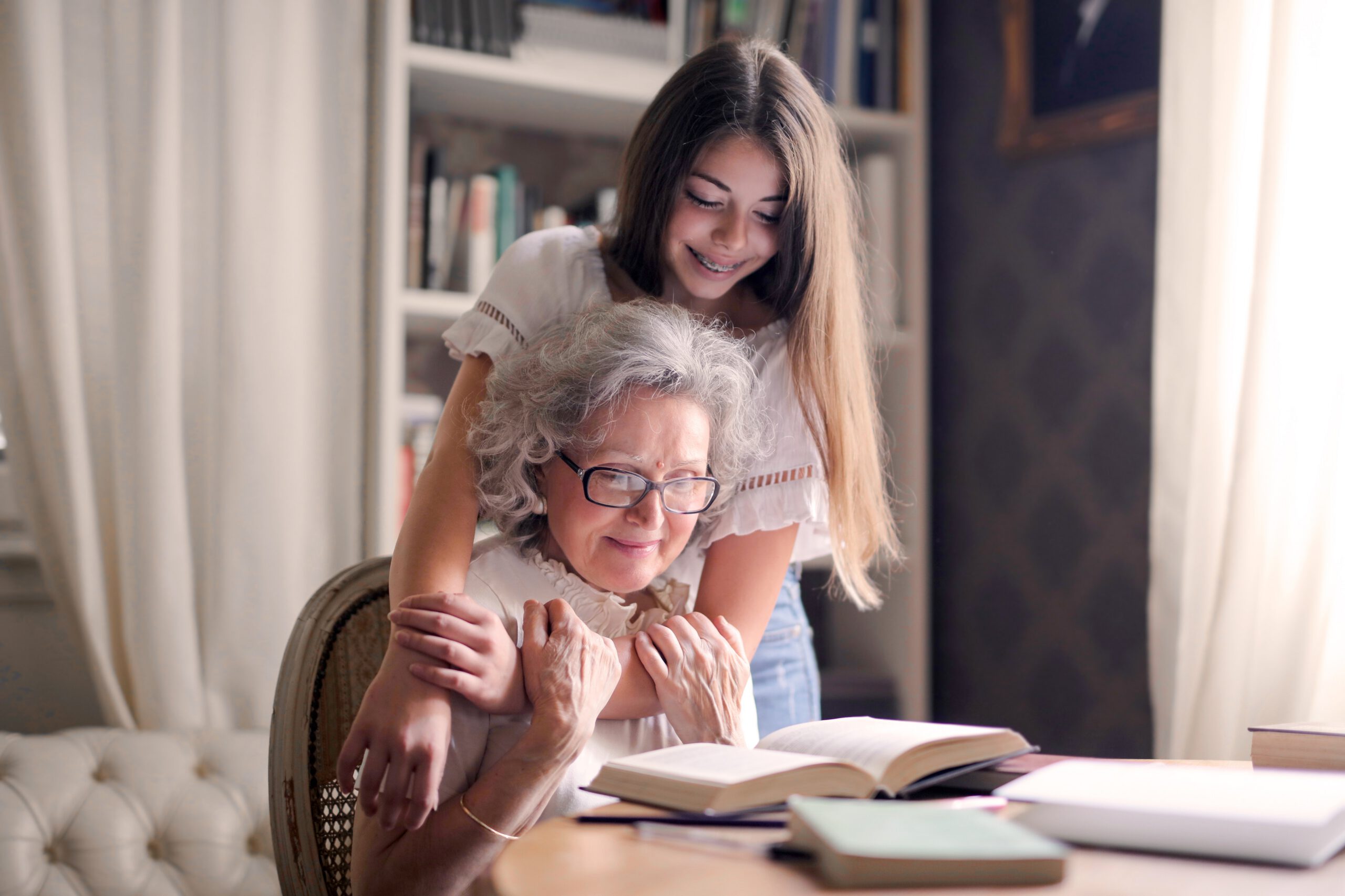 A smiling young woman hugs an elderly woman from behind as they sit at a table with an open book in a cozy, well-lit room filled with bookshelves.