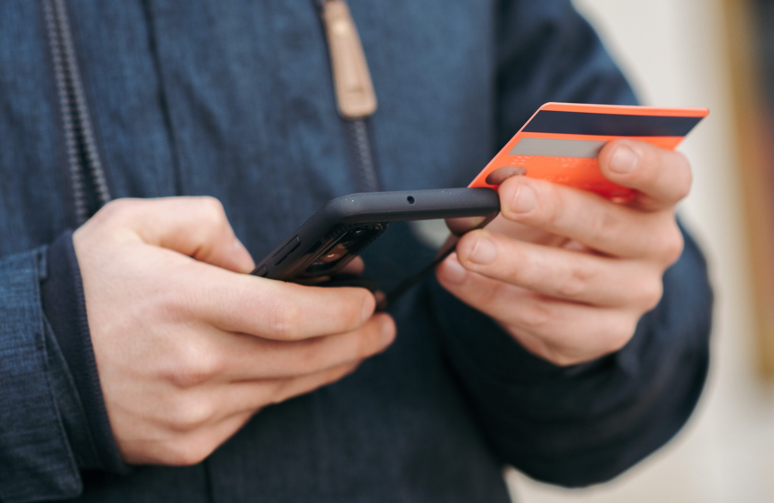 A person wearing a dark jacket holds a smartphone in one hand and an orange credit card in the other, appearing to make an online payment or check account details.
