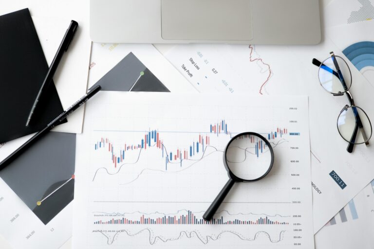A magnifying glass rests on a printed financial chart with candlestick patterns. Nearby are a laptop, pens, eyeglasses, and documents with graphs and data, suggesting data analysis or financial review.