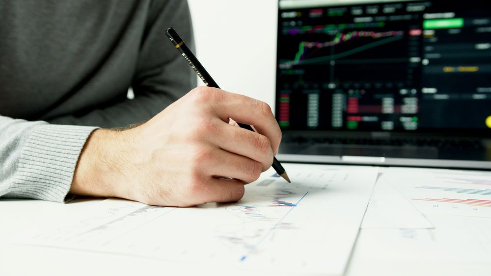 A person holding a pencil and analyzing printed financial charts on a desk, with a laptop displaying colorful stock market graphs and data in the background.