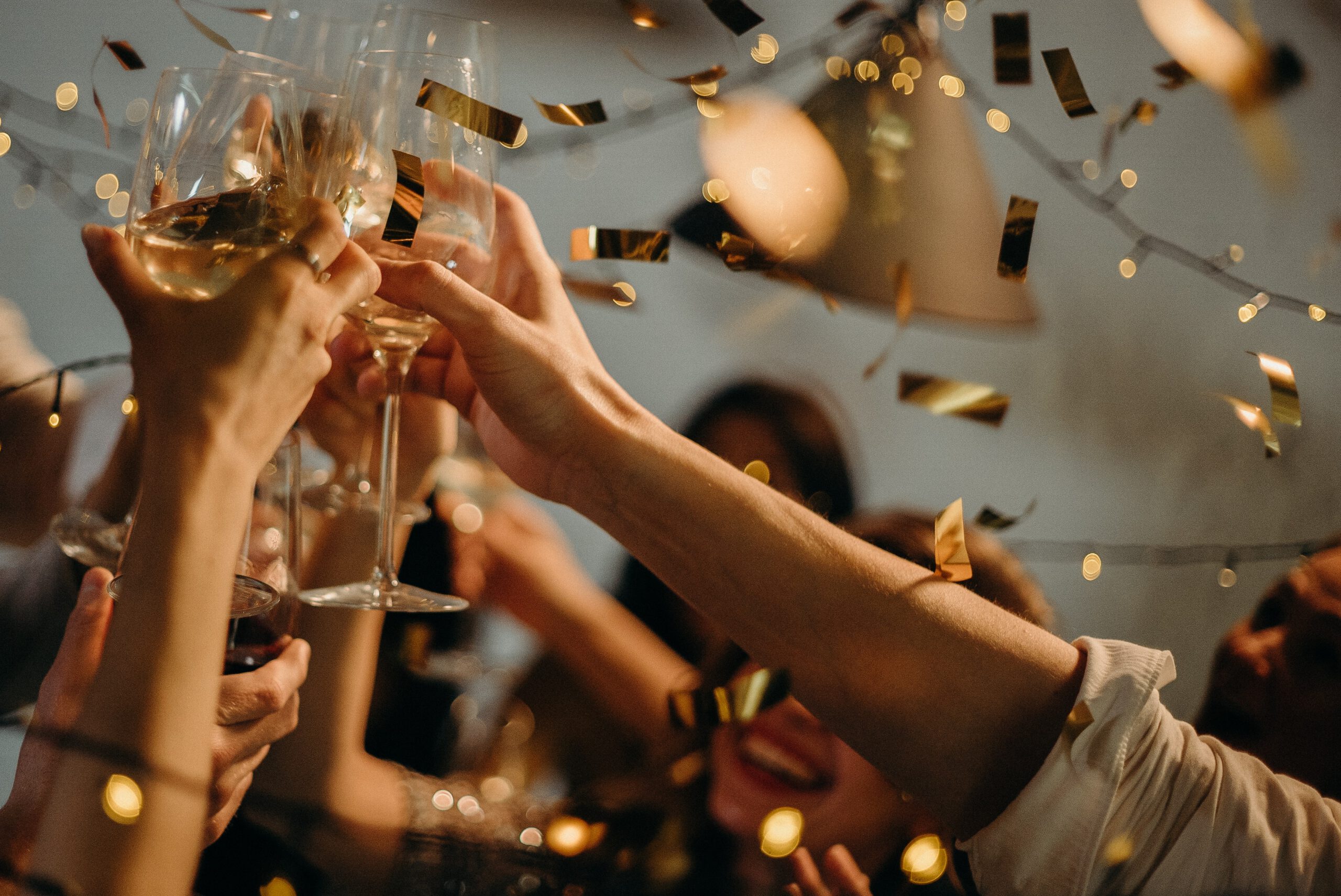 Hands raise glasses of champagne with gold confetti falling and fairy lights in the background.