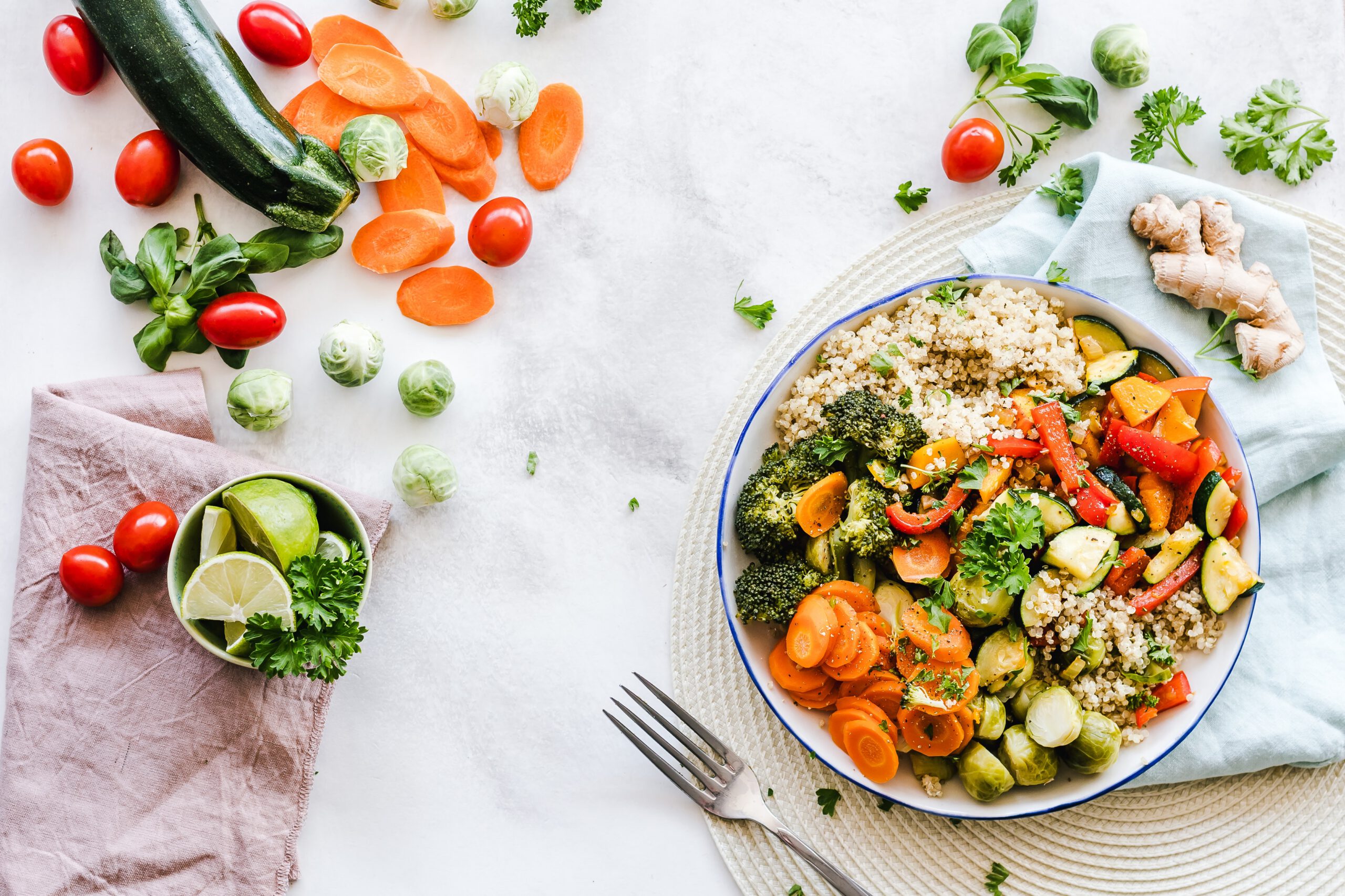A colorful bowl of quinoa topped with roasted vegetables, including carrots, broccoli, zucchini, and peppers, sits on a mat beside fresh veggies like tomatoes, carrot slices, a zucchini, ginger, limes, and leafy herbs.