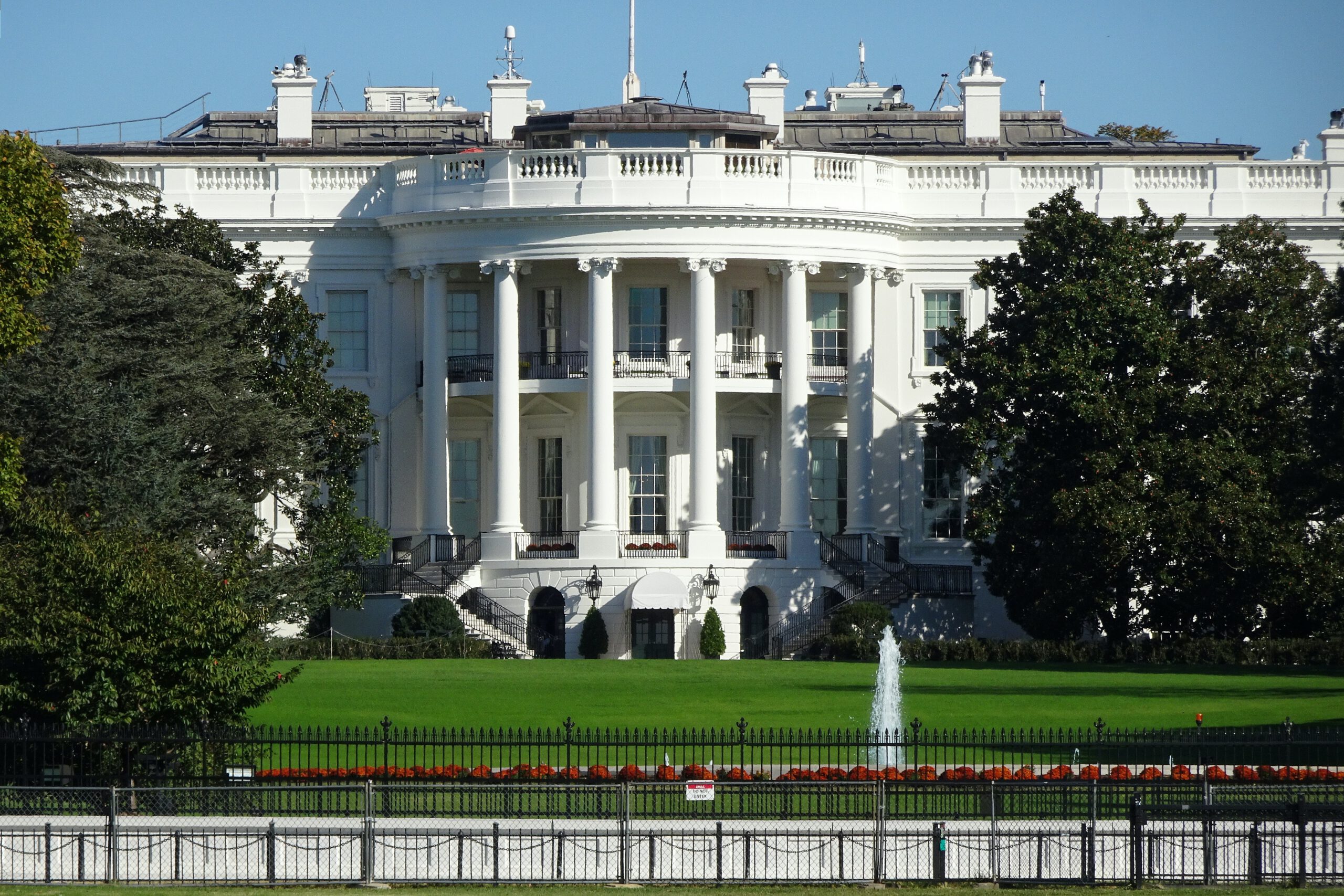 The White House, a large white neoclassical building with columns, is seen from the front surrounded by green lawns, trees, and a fountain under a clear blue sky.