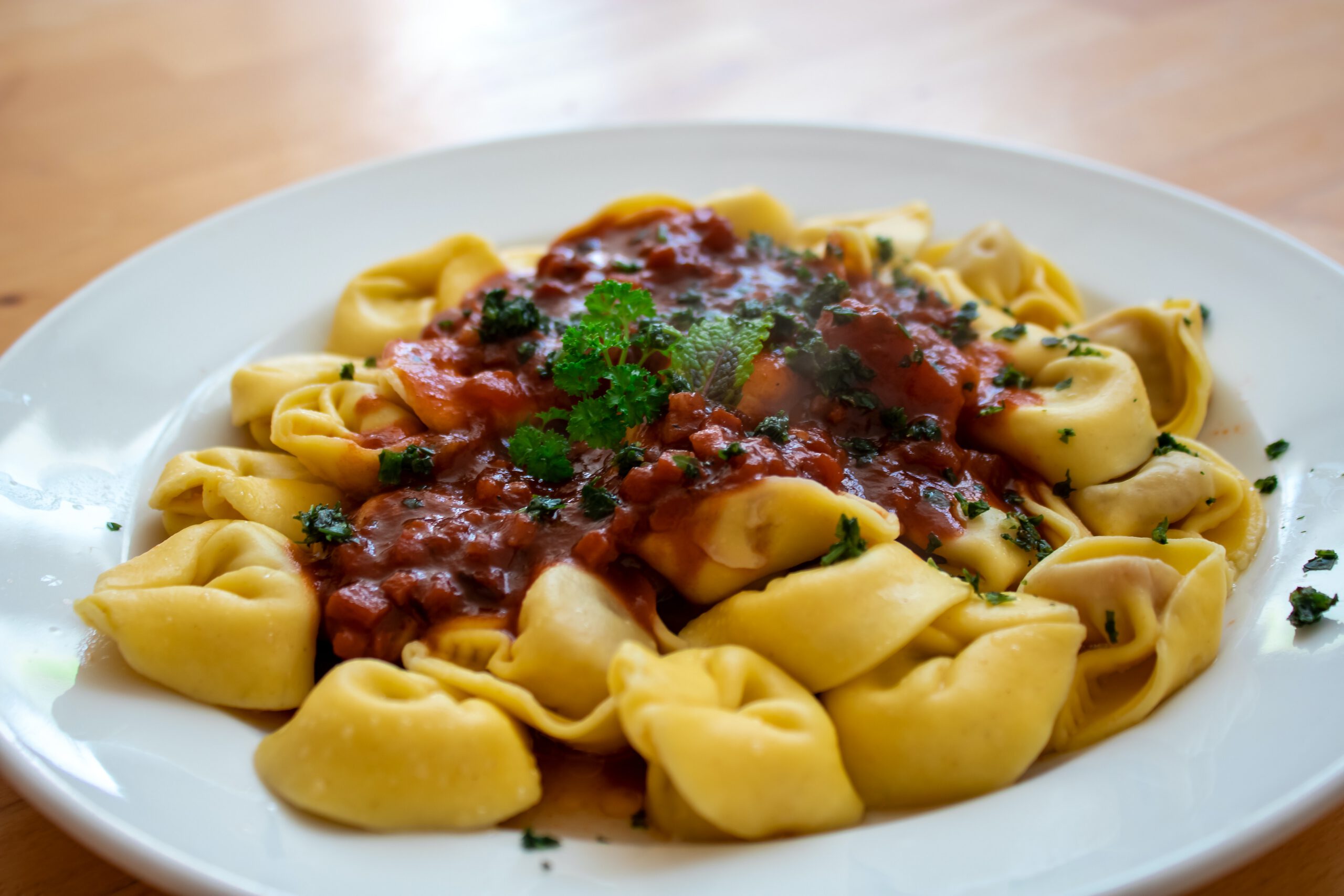 A plate of tortellini pasta arranged in a circle, topped with a chunky red tomato sauce and garnished with fresh parsley, served on a white plate on a wooden table.