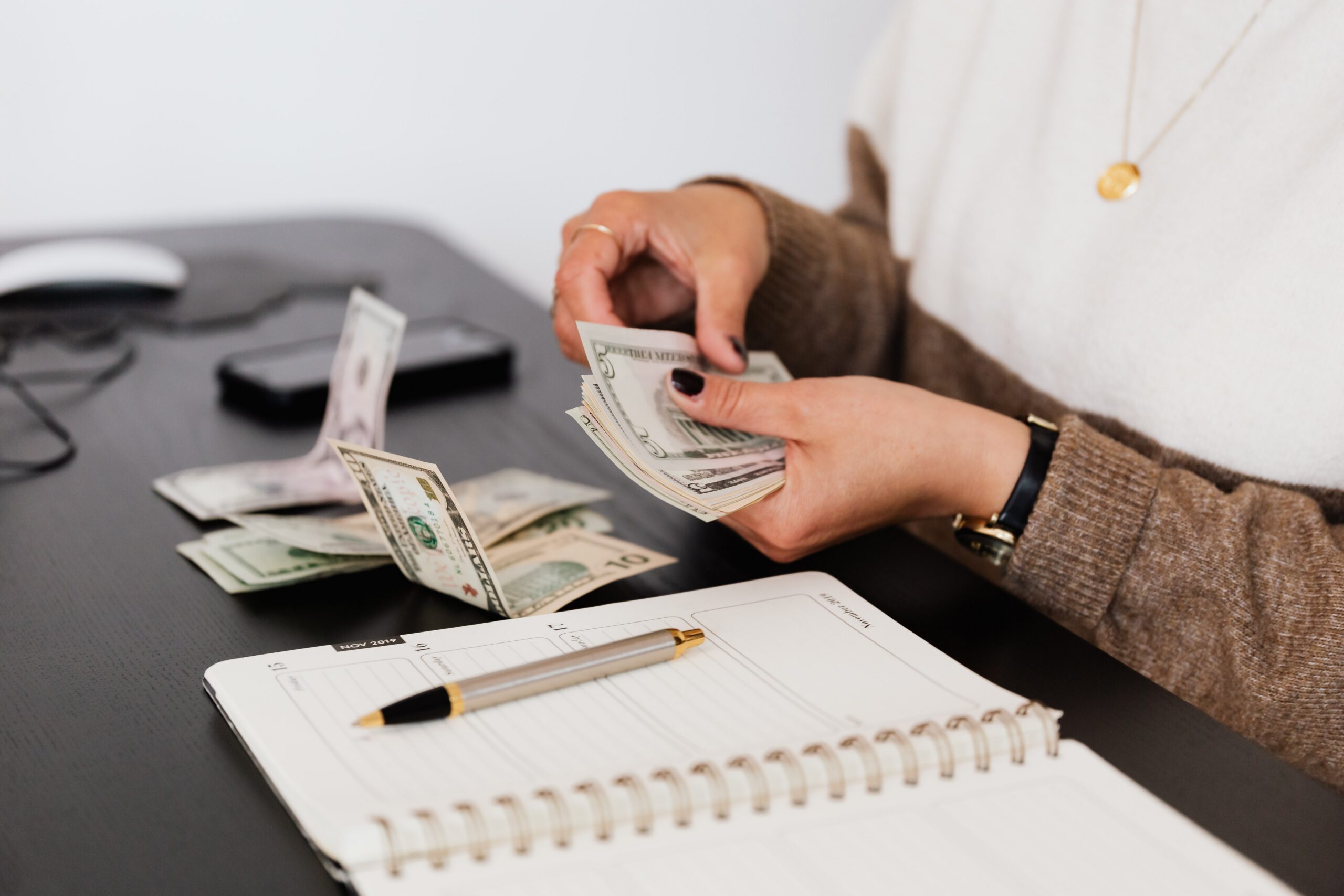 A person wearing a brown and white sweater counts US dollar bills at a black desk with a notebook, pen, and smartphone nearby.