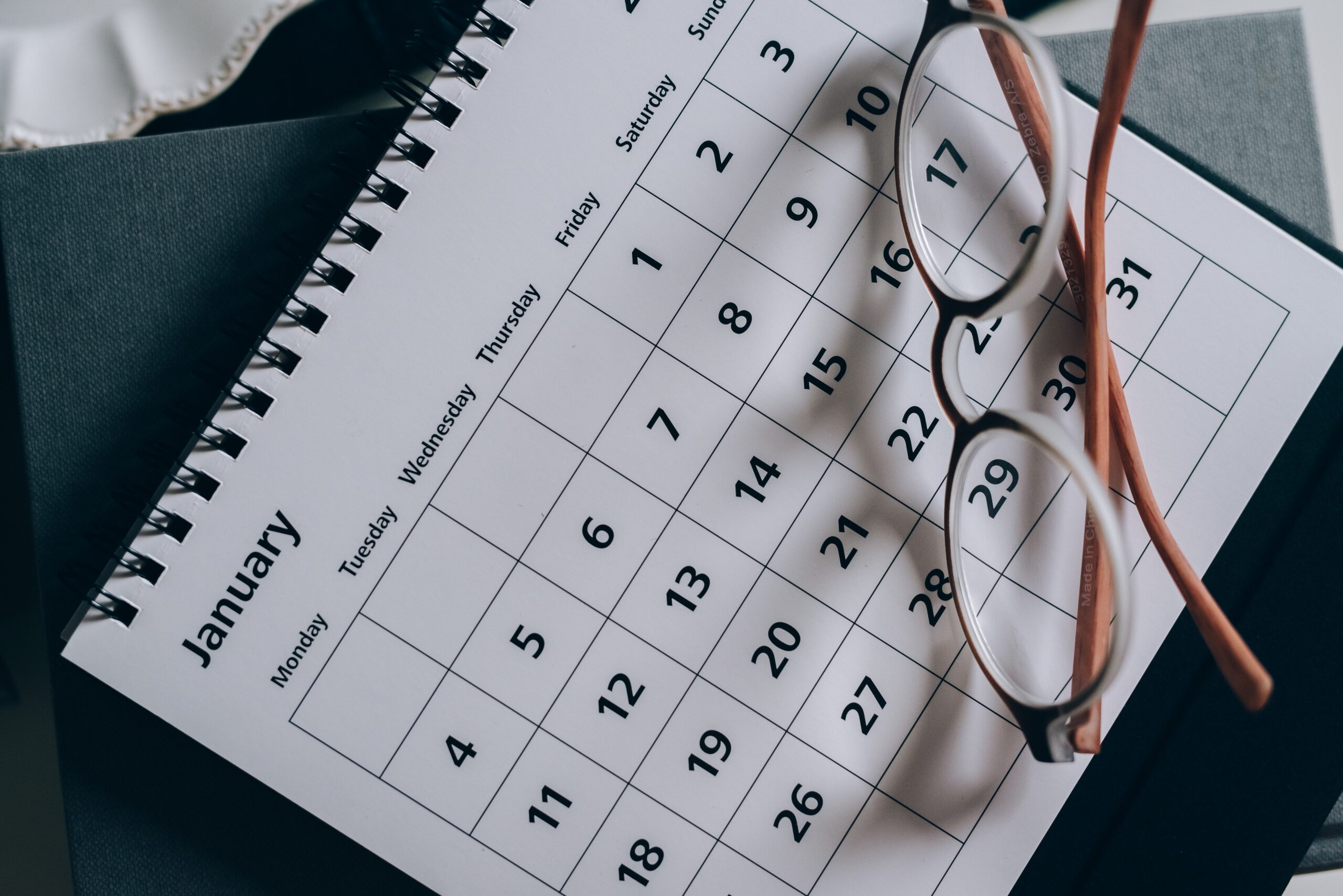 A pair of eyeglasses rests on an open spiral-bound calendar displaying the month of January, with dates clearly visible. The calendar sits on a stack of books or notebooks.