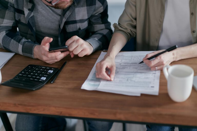 Two people sit at a wooden table, reviewing documents and filling out forms with pens. A calculator, a smartphone, and a white mug are also on the table.