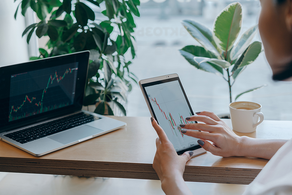 A person uses a tablet to analyze stock charts beside a laptop displaying similar charts. A coffee cup and green plants are on the wooden desk by a window.