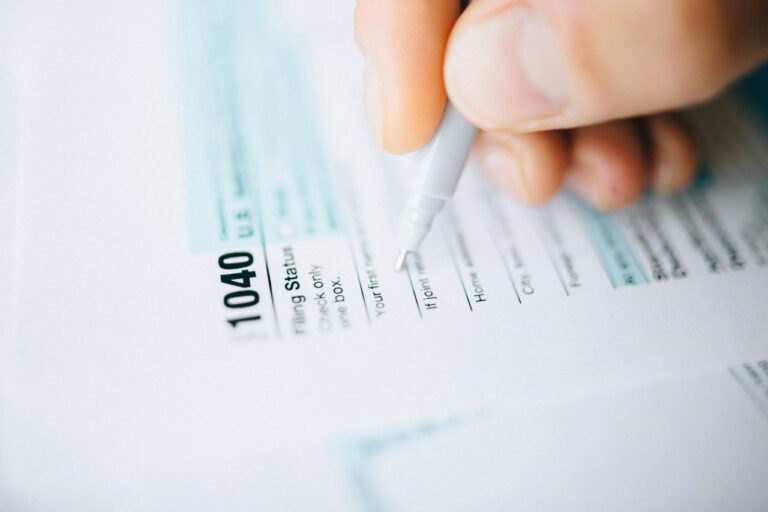A close-up of a person’s hand holding a pen while filling out a U.S. 1040 tax form, with the forms text and sections partially visible.