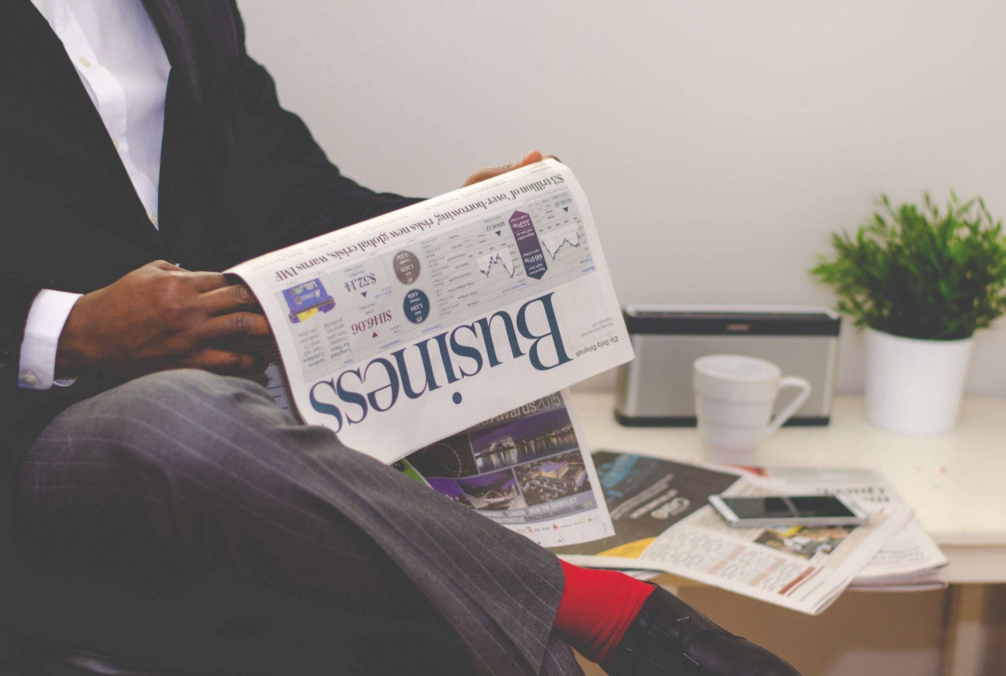 A person in a suit with red socks reads a business newspaper in an office setting, with a coffee cup, a tablet, papers, and a potted plant on a white table nearby.