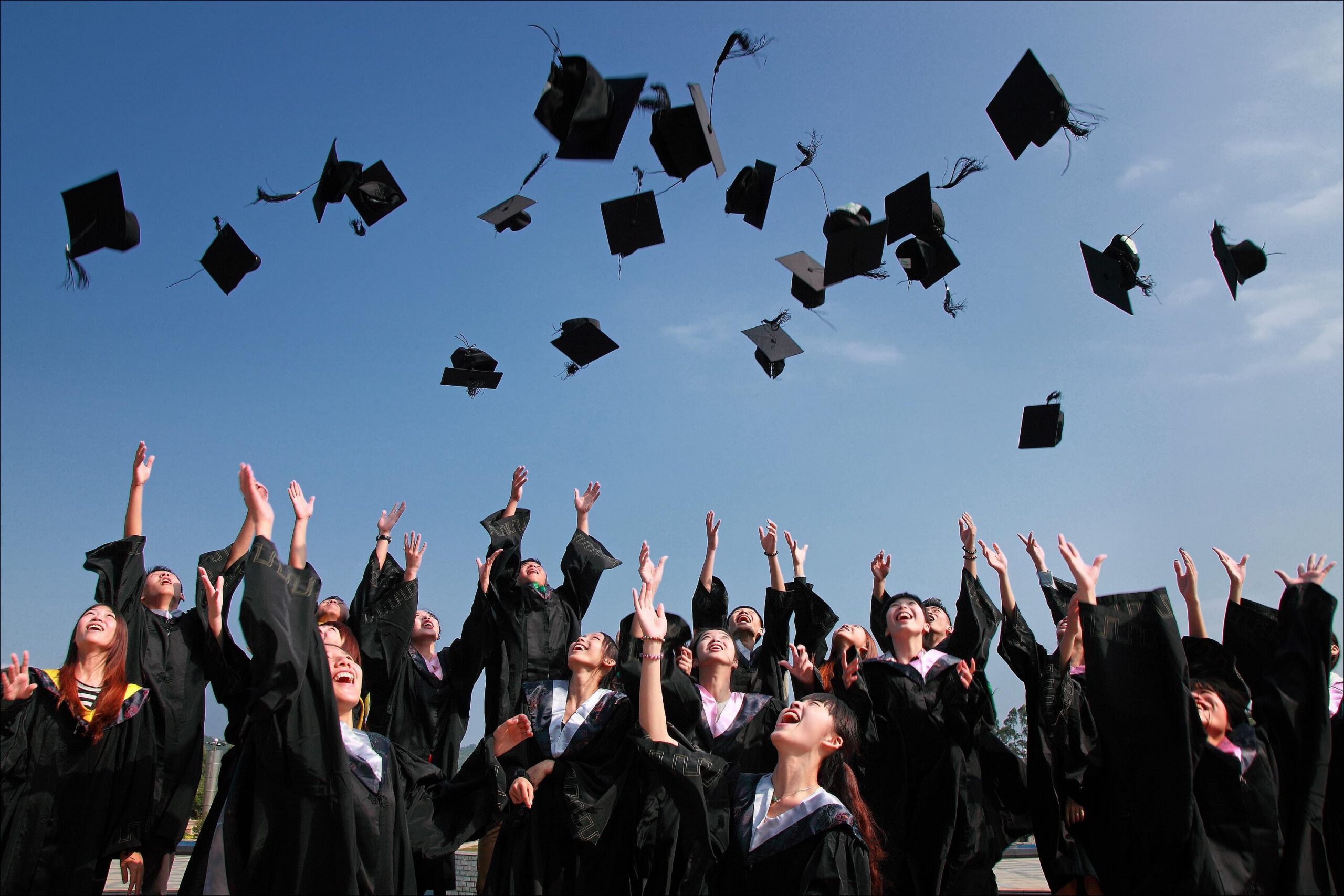 A group of graduates in black caps and gowns celebrate by tossing their caps into the air under a clear blue sky.