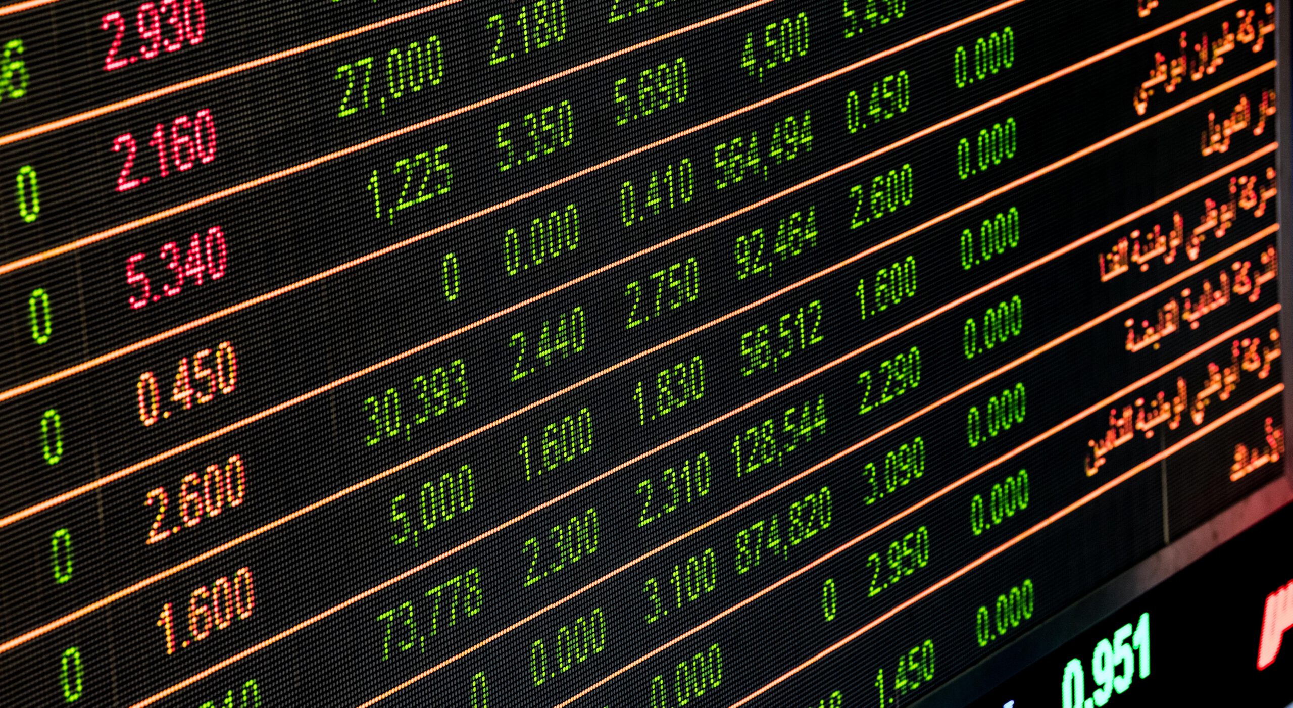 A digital display shows rows of stock market data with numbers in green, red, and white, and some Arabic text on a black background. The numbers indicate prices, volumes, and market changes.