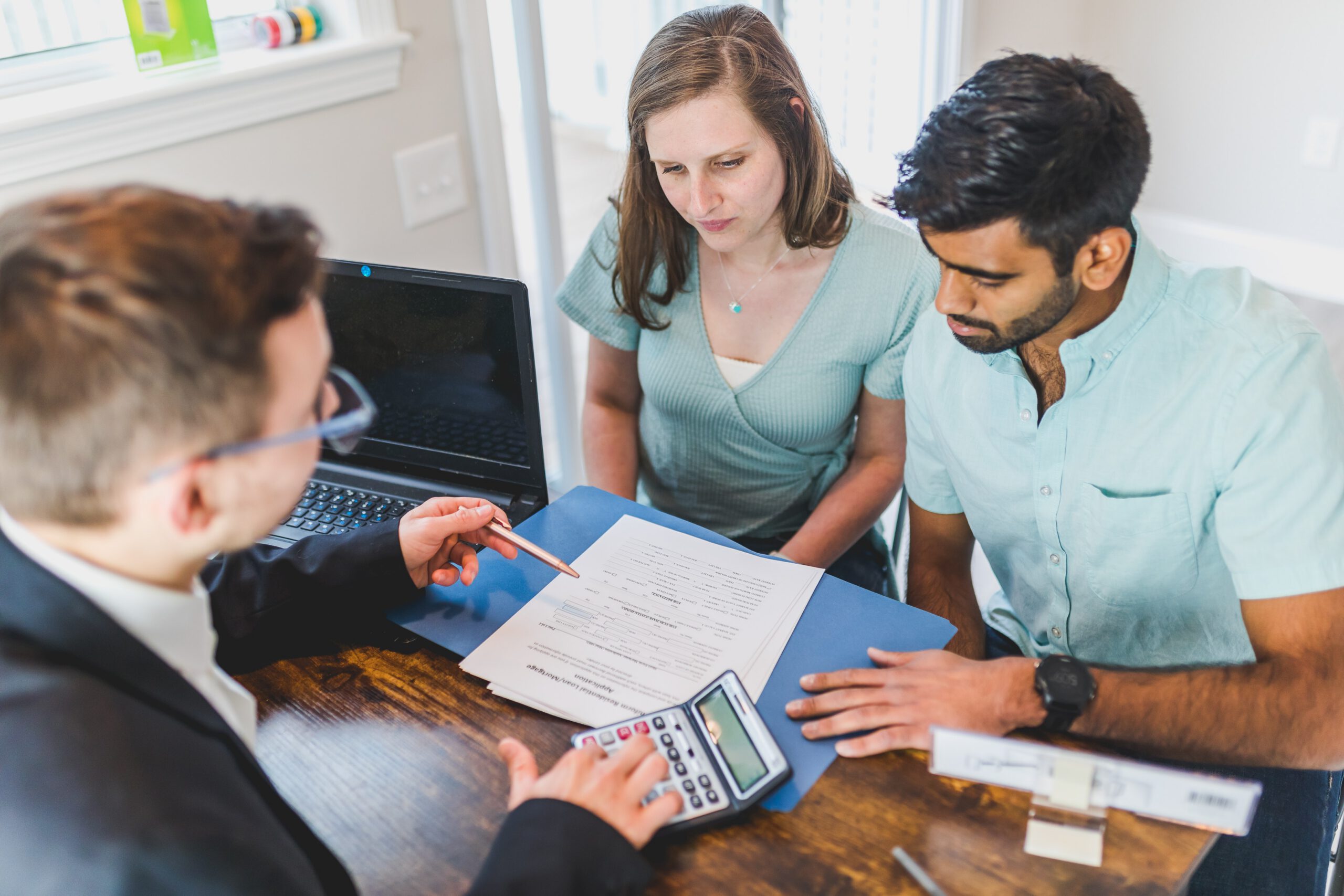 A couple sits at a desk with a professional, reviewing documents and using a calculator, with a laptop nearby, suggesting a financial or legal consultation.