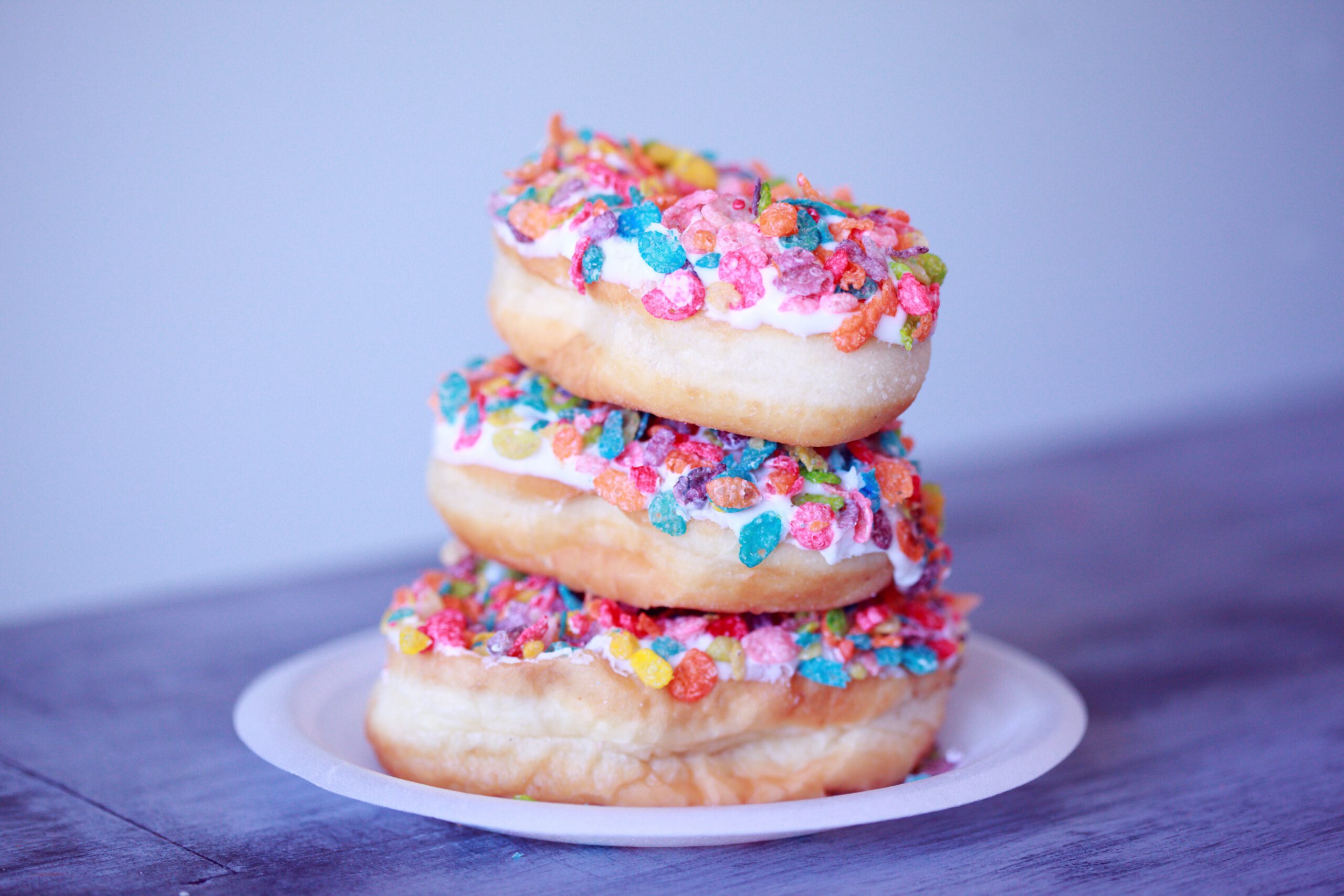 Three donuts stacked on a white plate, topped with white icing and colorful cereal pieces, against a light blue background.