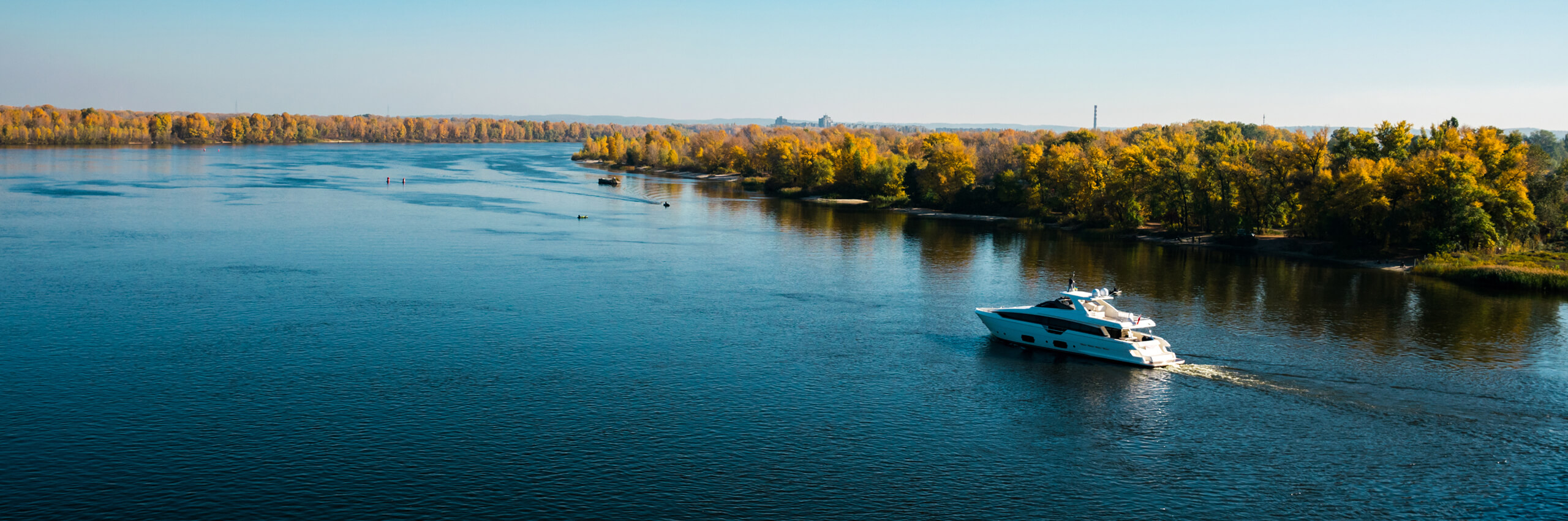 A white yacht cruises on a wide, calm river with a shoreline of trees in autumn colors under a clear blue sky.