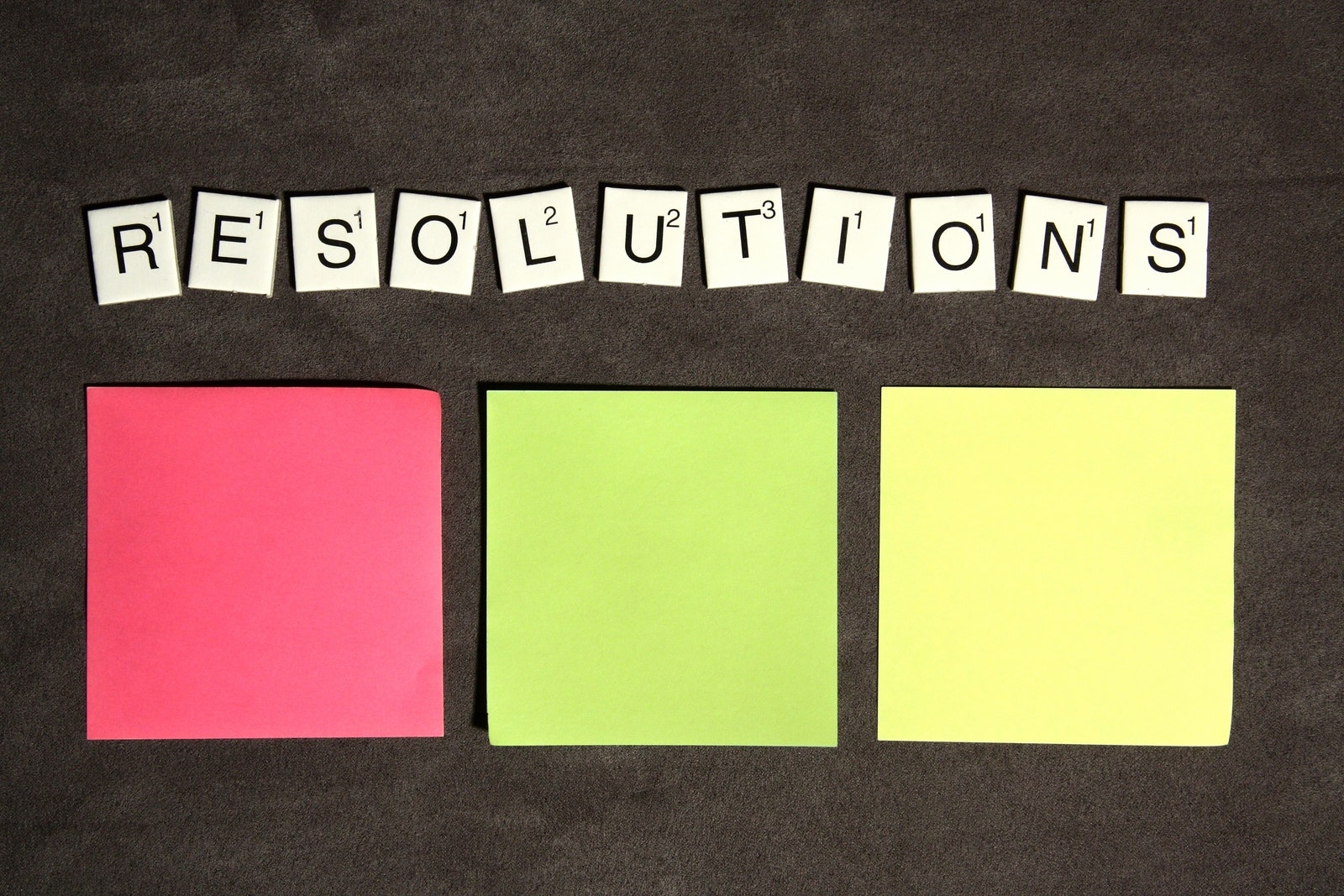 Three colorful sticky notes (pink, green, yellow) are arranged in a row below Scrabble tiles spelling RESOLUTIONS on a dark surface.