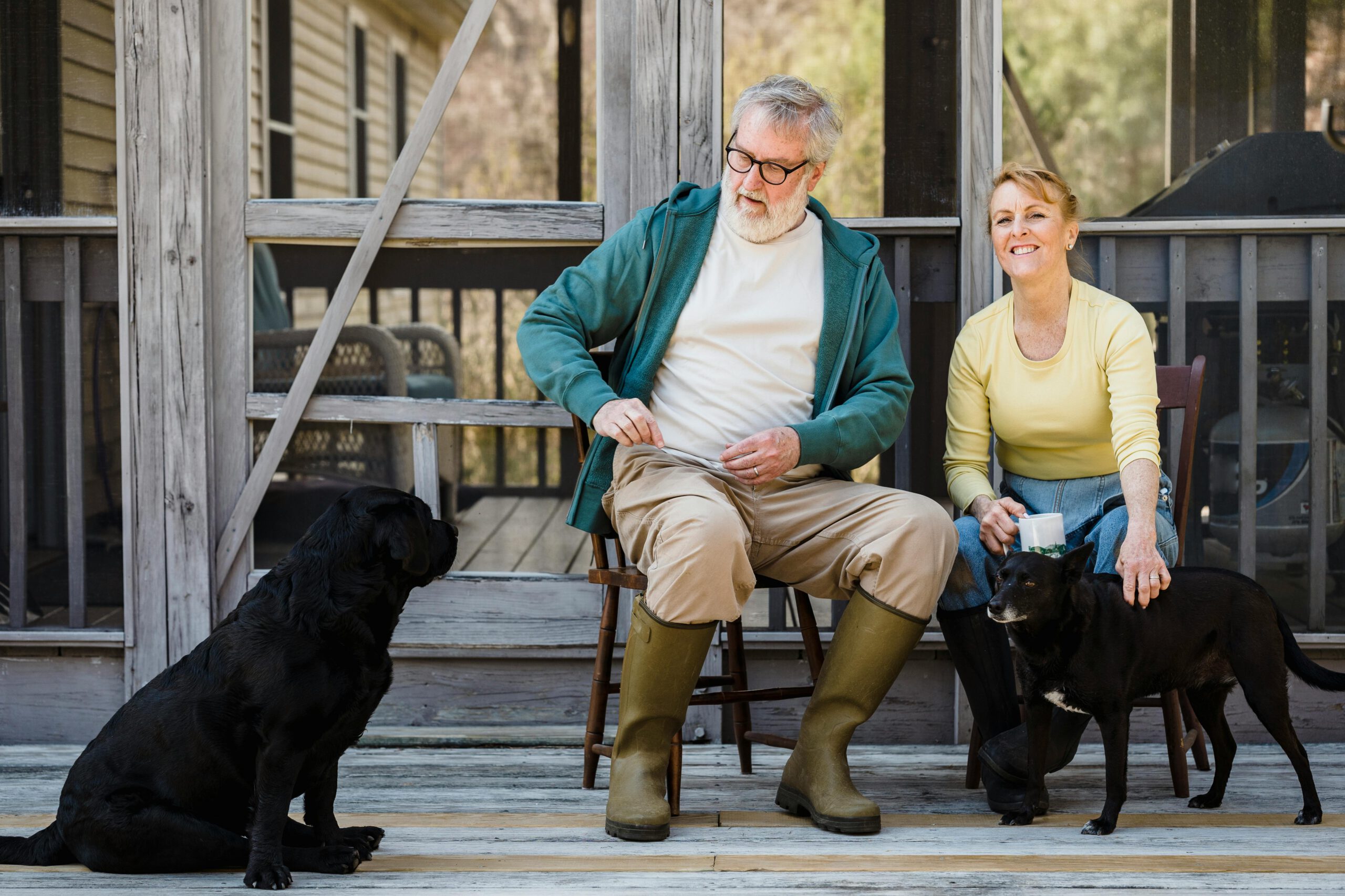 An older man and woman sit on a wooden porch with two black dogs; the man is wearing boots and a green jacket, while the woman is petting one dog. Both appear content and are enjoying time outdoors.